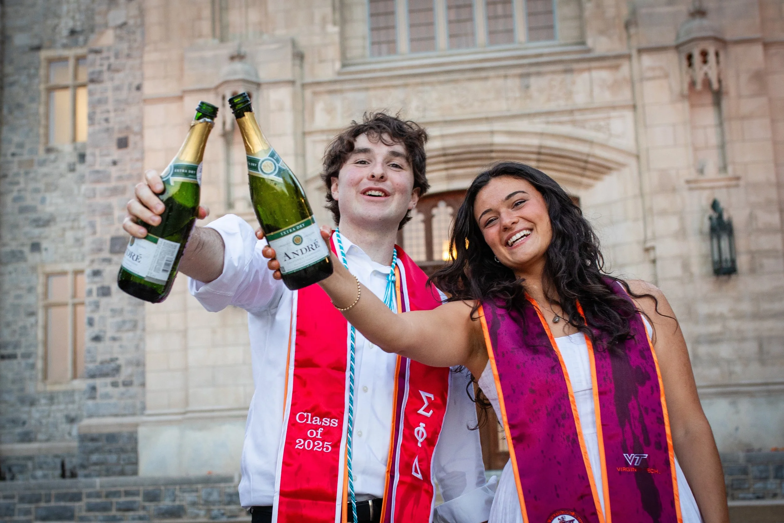 Two graduates celebrate outdoors, holding bottles of champagne, with a historic stone building in the background. The male graduate wears a white shirt and a red stole with 'Class of 2025' and Greek letters, while the female graduate wears a white dr