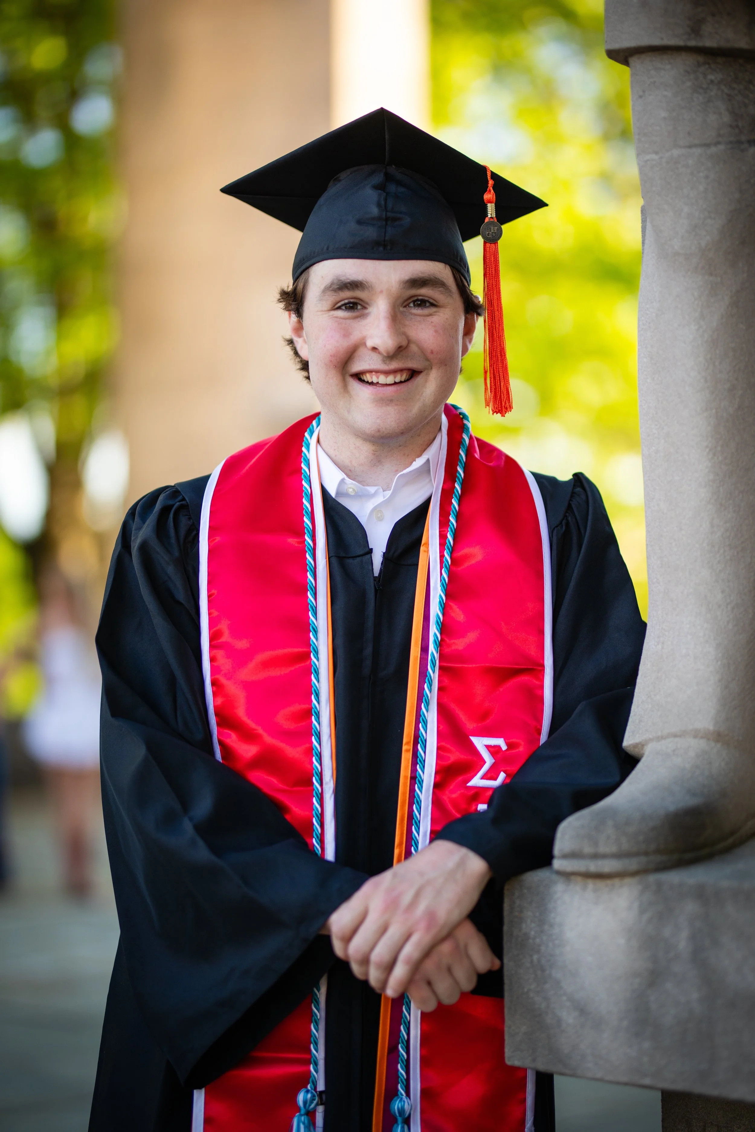 A young man in a graduation cap and gown, with honor cords and a red stole, smiles and leans against a stone monument outdoors with green blurred trees in the background.