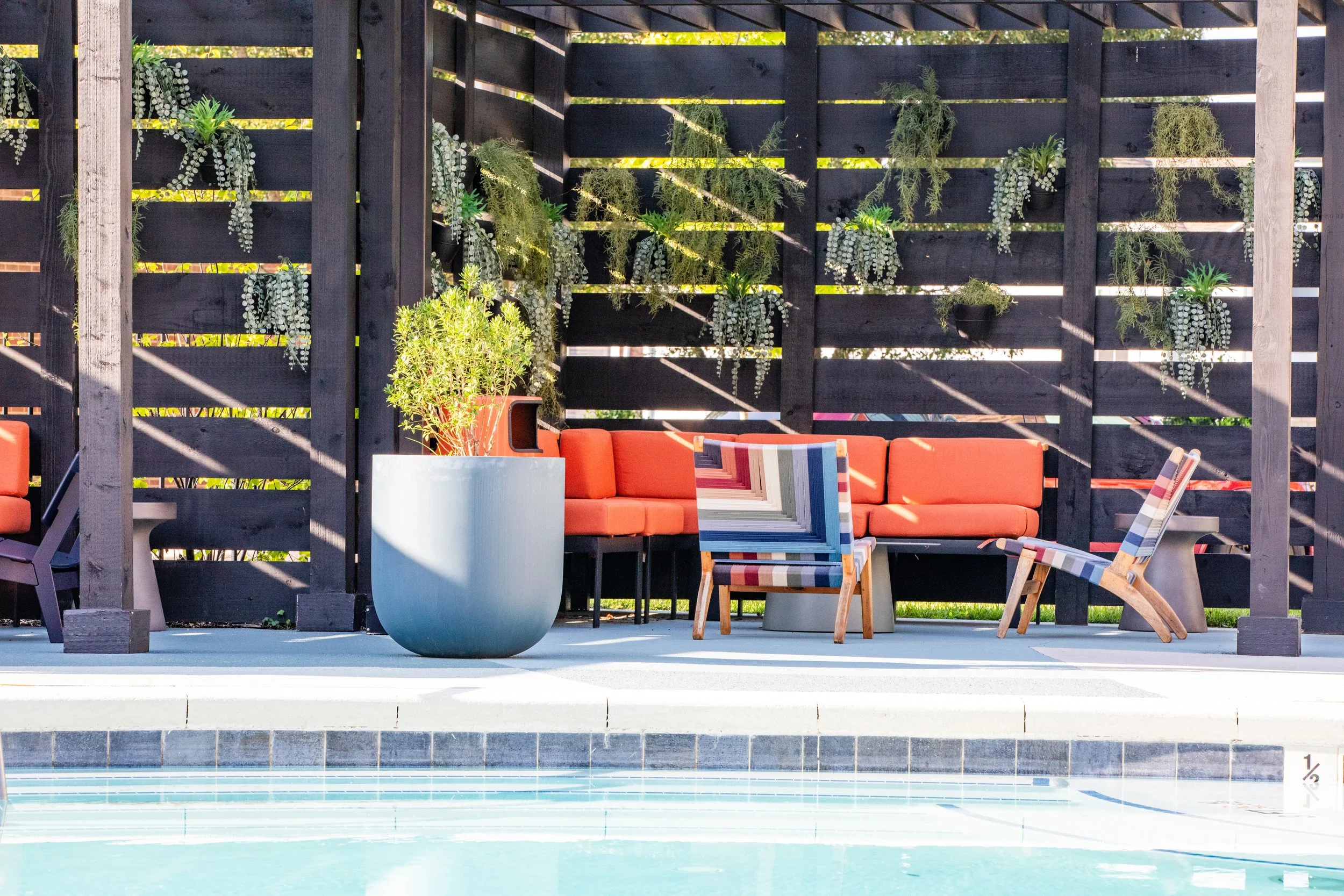 Outdoor poolside seating area with orange cushioned bench, colorful striped chairs, potted plant, and a dark wooden privacy fence with hanging plants.
