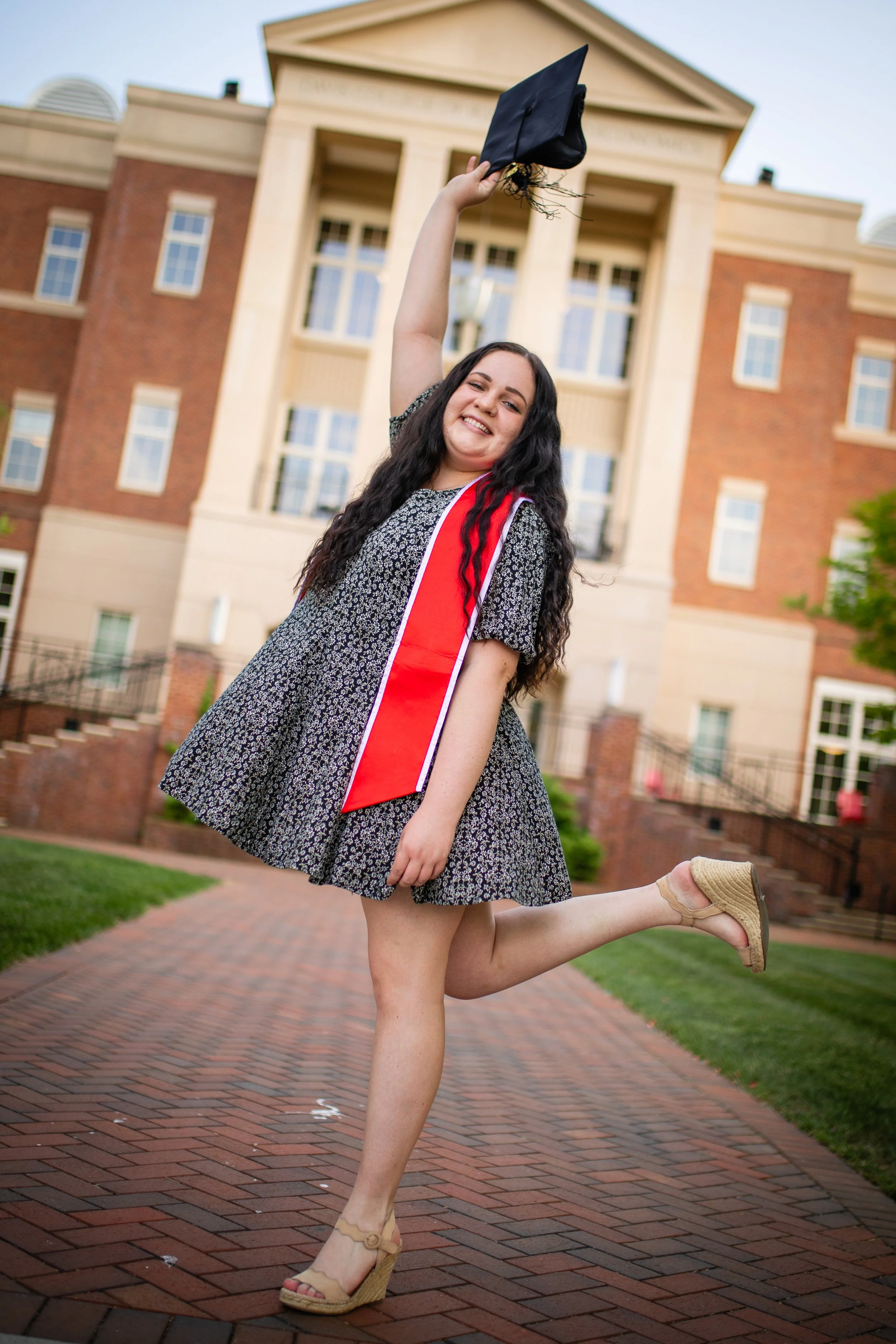 A young woman in a dress with a red honor cord celebrates her graduation outdoors in front of a large building, holding a graduation cap in the air and smiling.