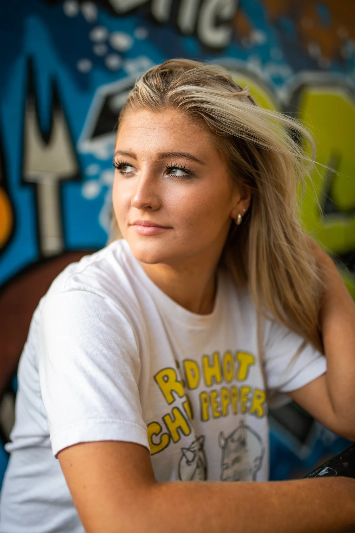 A young woman with blonde hair looking to the side, wearing a white t-shirt with yellow and black lettering and graphics, sitting outdoors near a colorful graffiti wall.