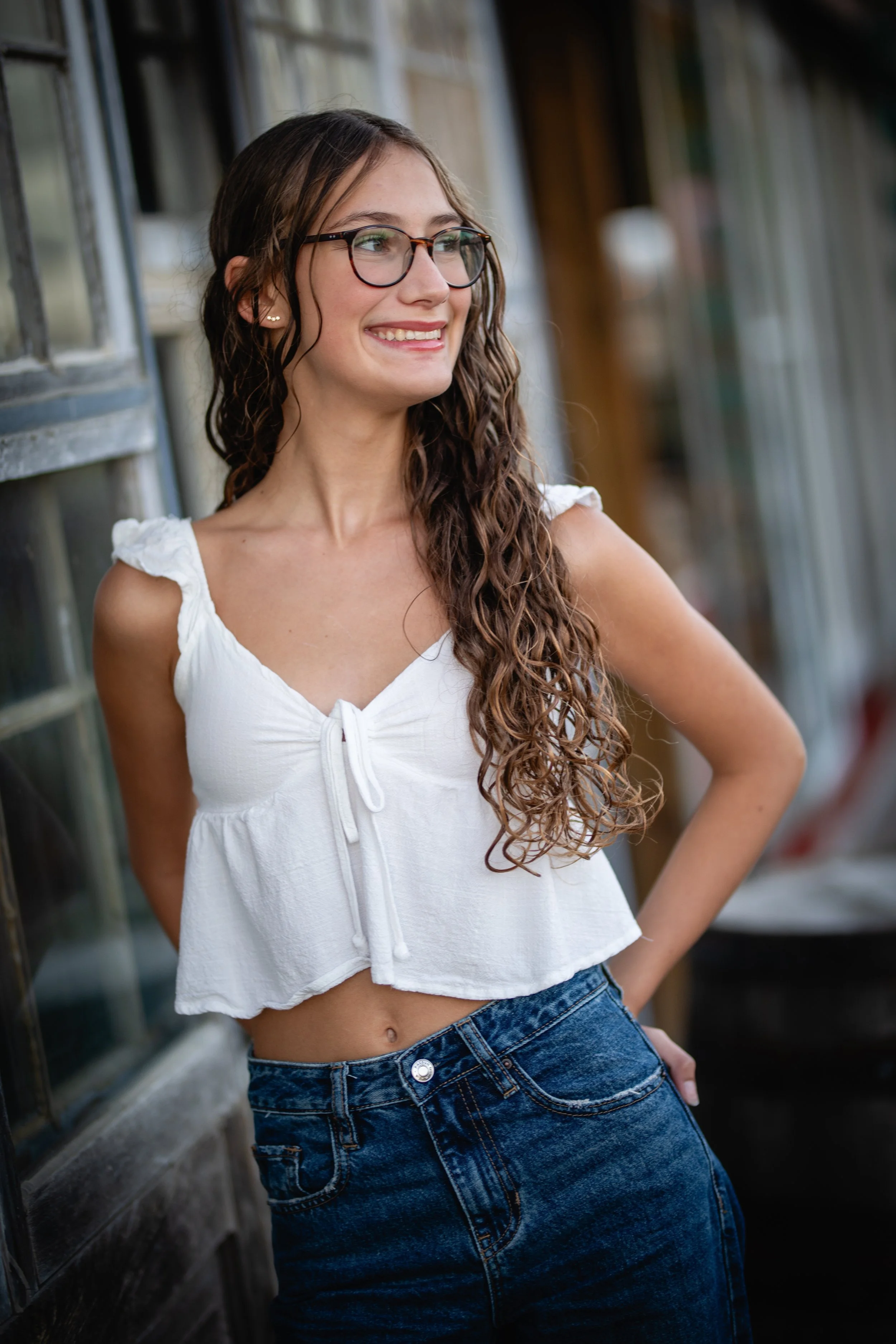 A young woman with long, curly brown hair and glasses, smiling, outdoors at a rustic location with wooden and glass structures, wearing a white sleeveless top and dark blue jeans.