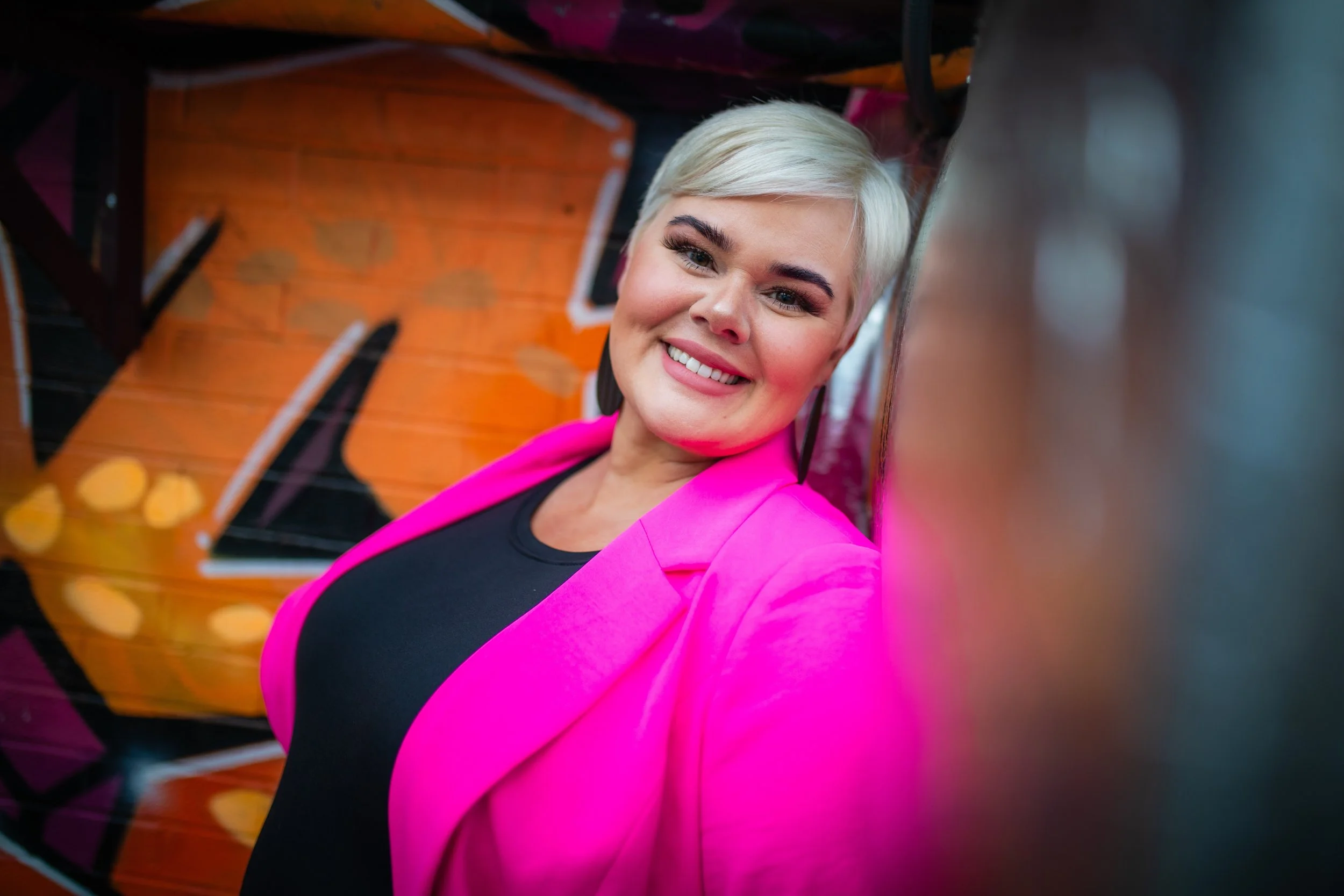 A smiling woman with short platinum blonde hair, wearing a bright pink blazer over a black top, leaning against a wall with colorful graffiti art.