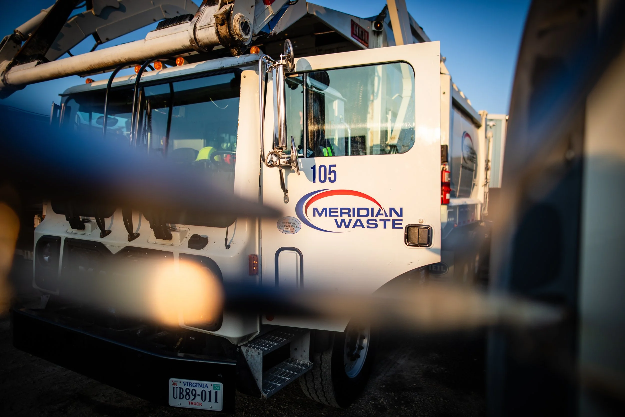 A Meridian Waste truck parked behind a chain-link fence, with the company's logo on the side and Virginia license plate visible.
