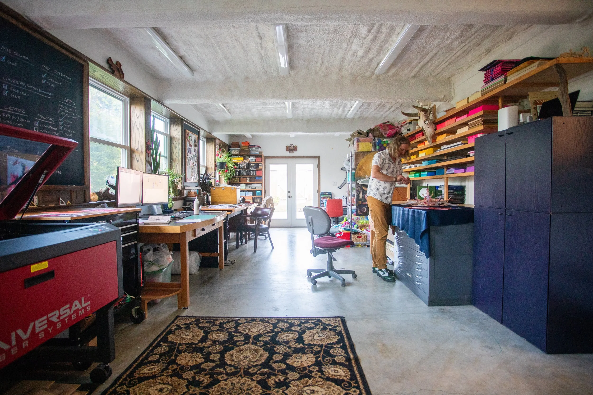 A spacious, bright studio with a woman working amidst shelves filled with colorful craft supplies and decorations. Multiple workstations with computers and plants are along the left side, and there's an area with a blackboard and a rug on the floor.