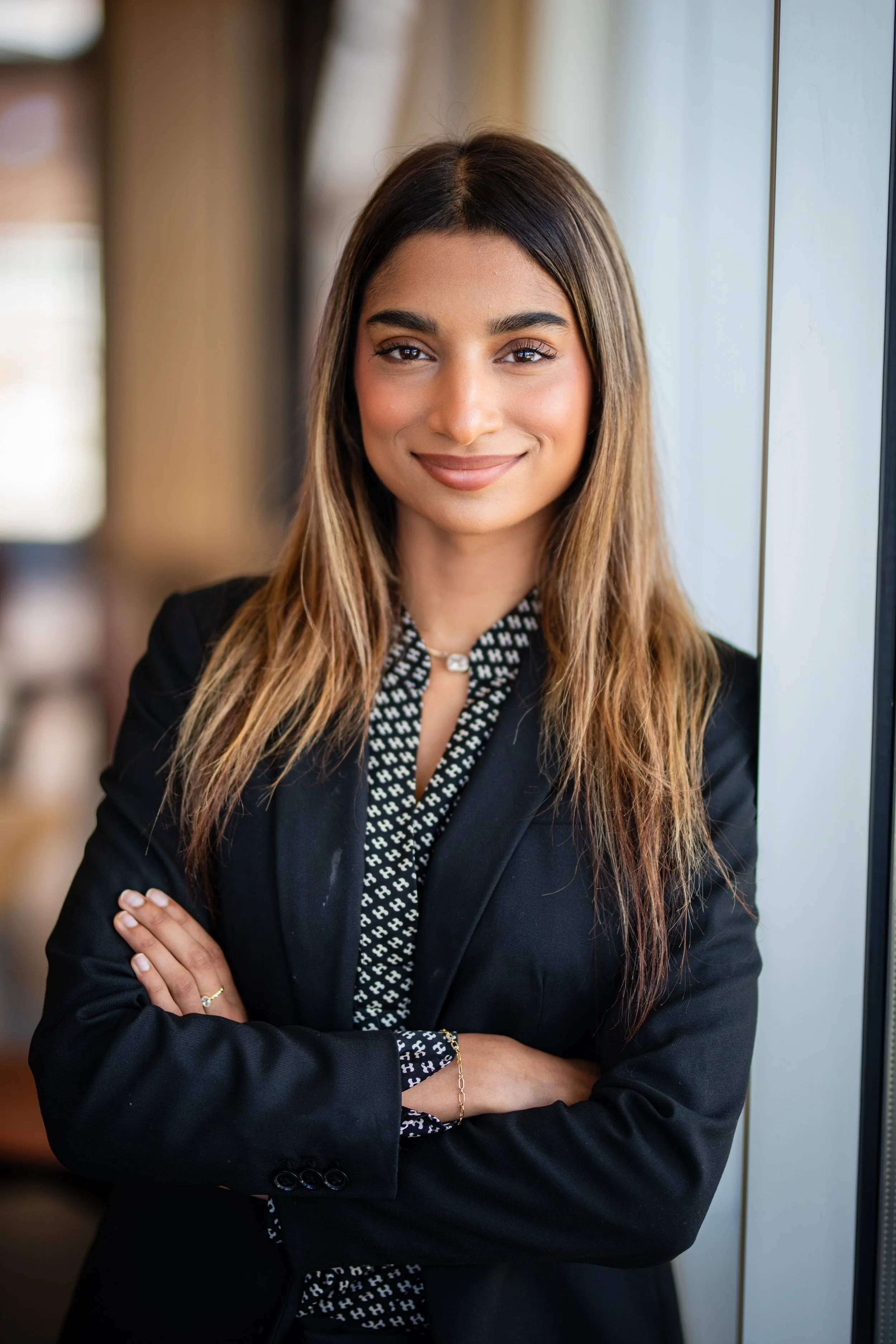A young woman with long brown hair and light skin, wearing a black blazer and patterned blouse, smiling and crossing her arms in front of a window in a professional setting.