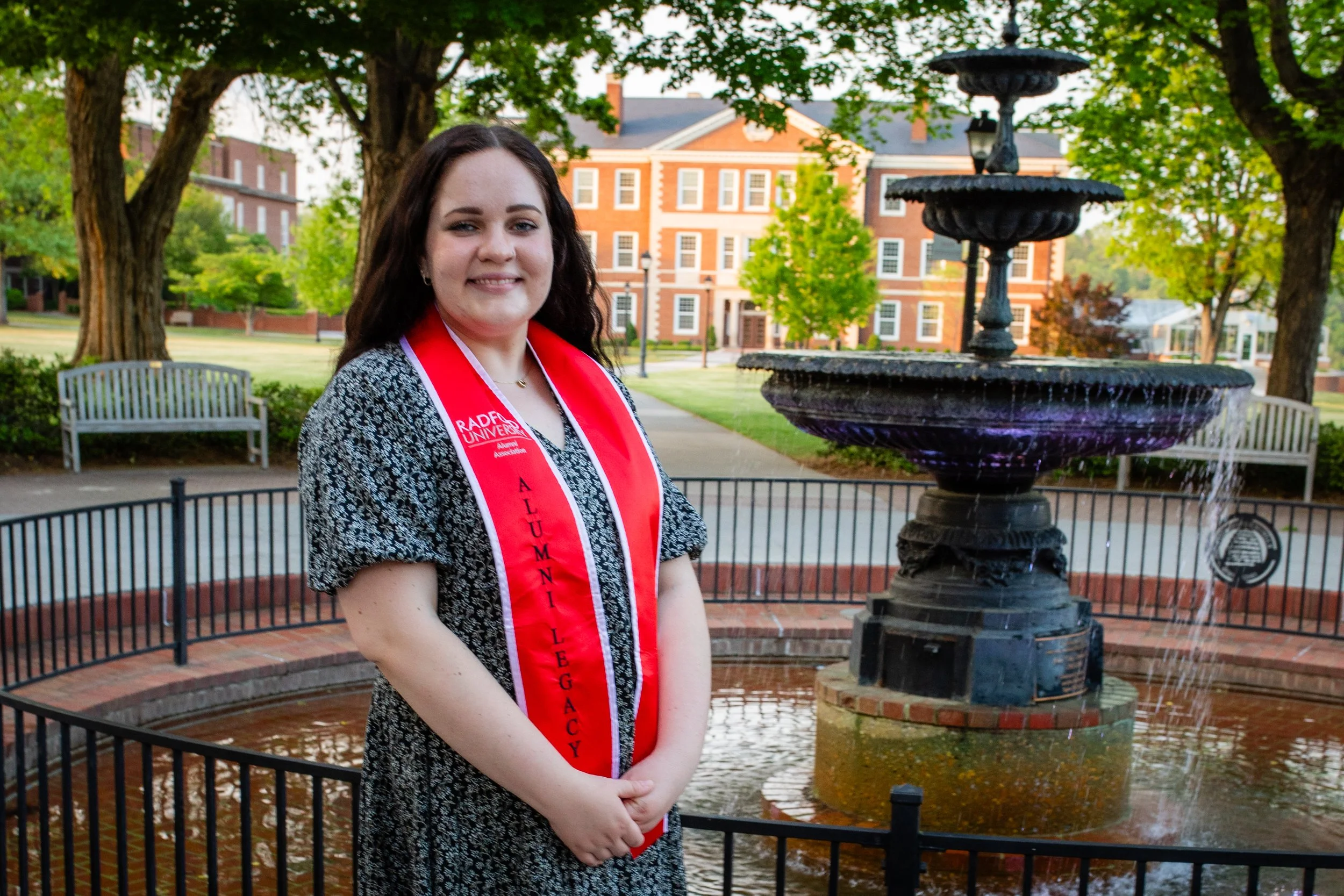 Young woman in a black and white patterned dress with a red graduation stole standing in front of a fountain on a college campus.