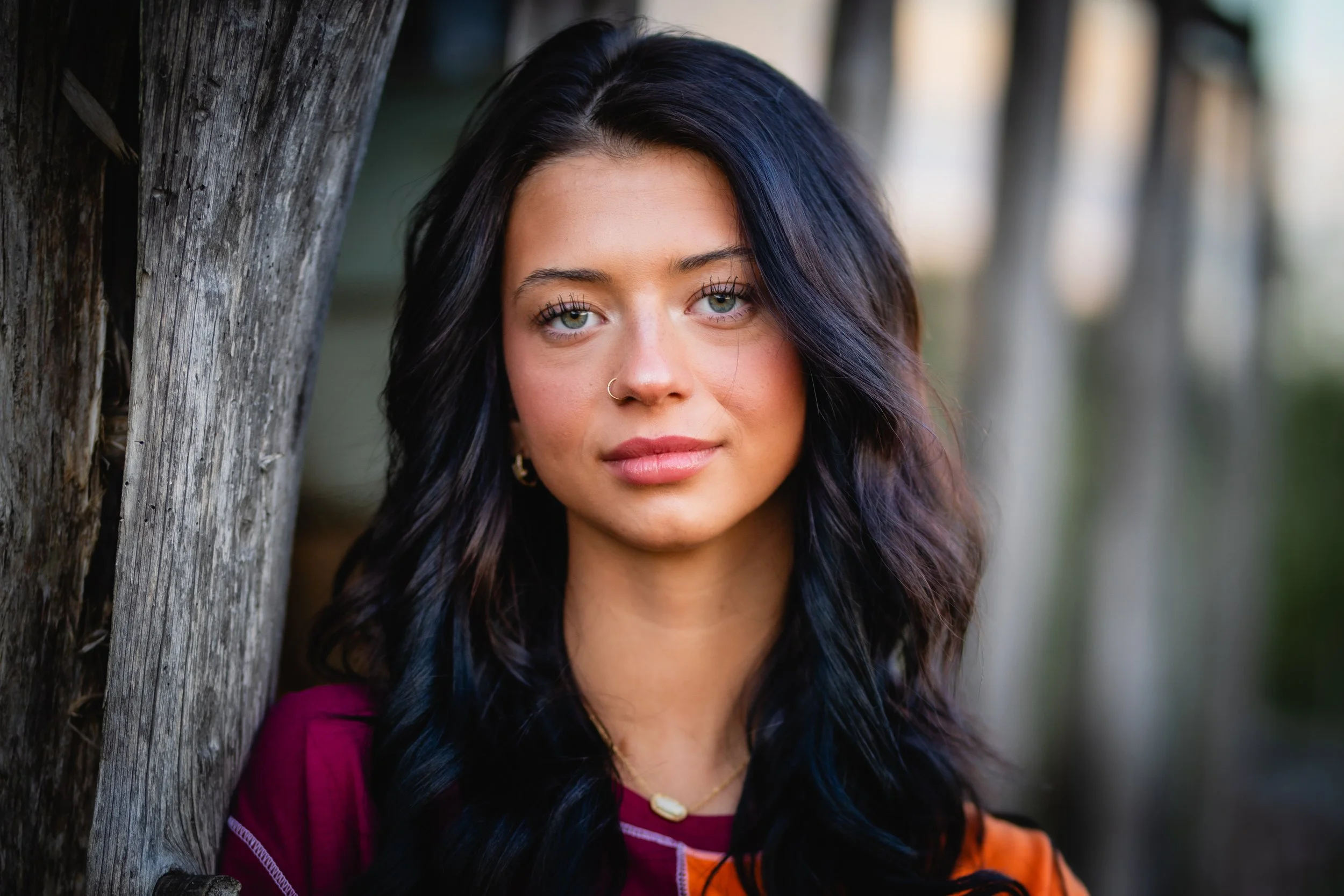 Close-up portrait of a young woman with dark, wavy hair, wearing a nose ring and jewelry, standing outdoors near a weathered wooden fence.