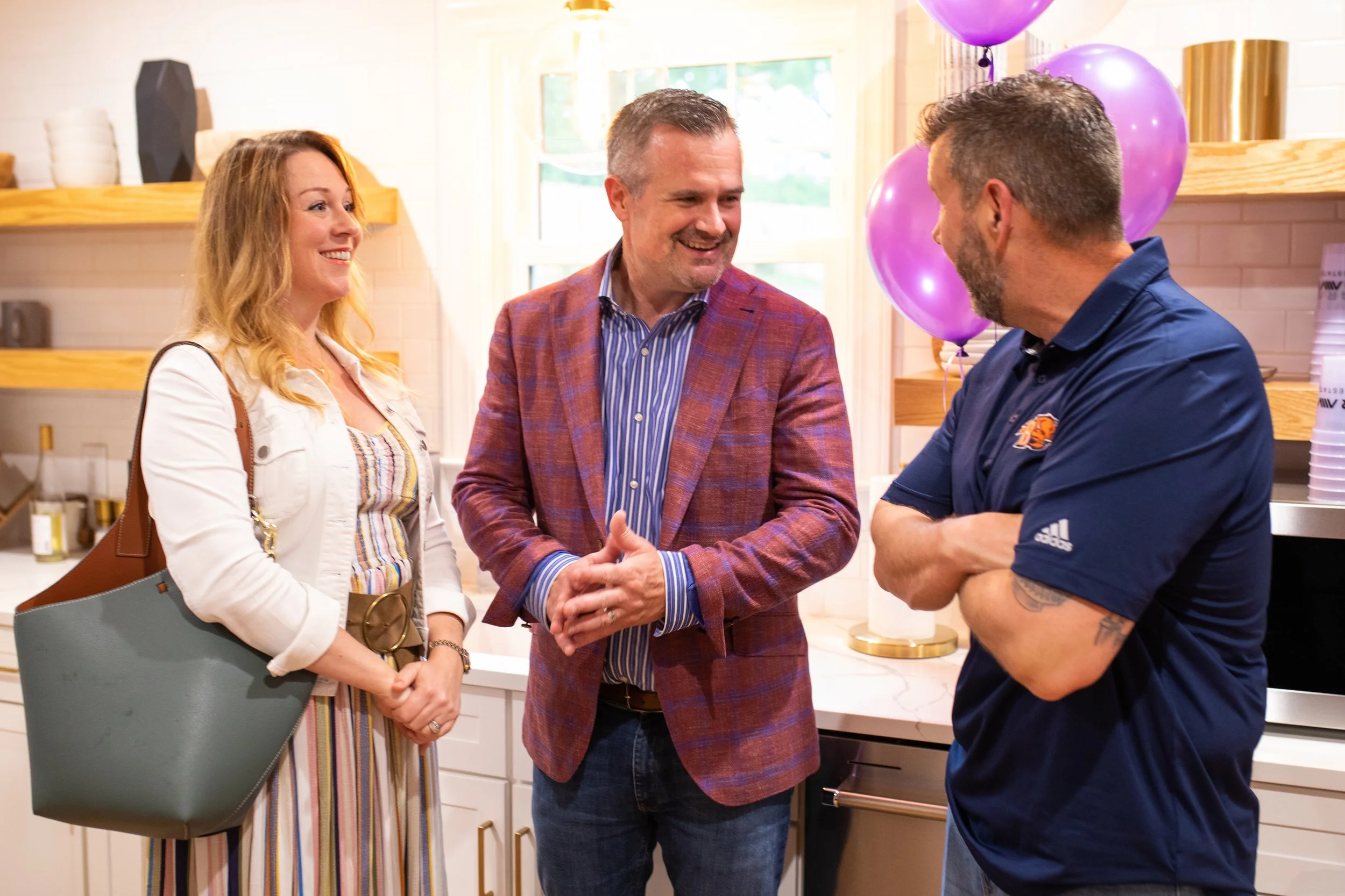 Three people, two men and a woman, are talking and smiling at each other at a party in a bright kitchen with purple balloons in the background.