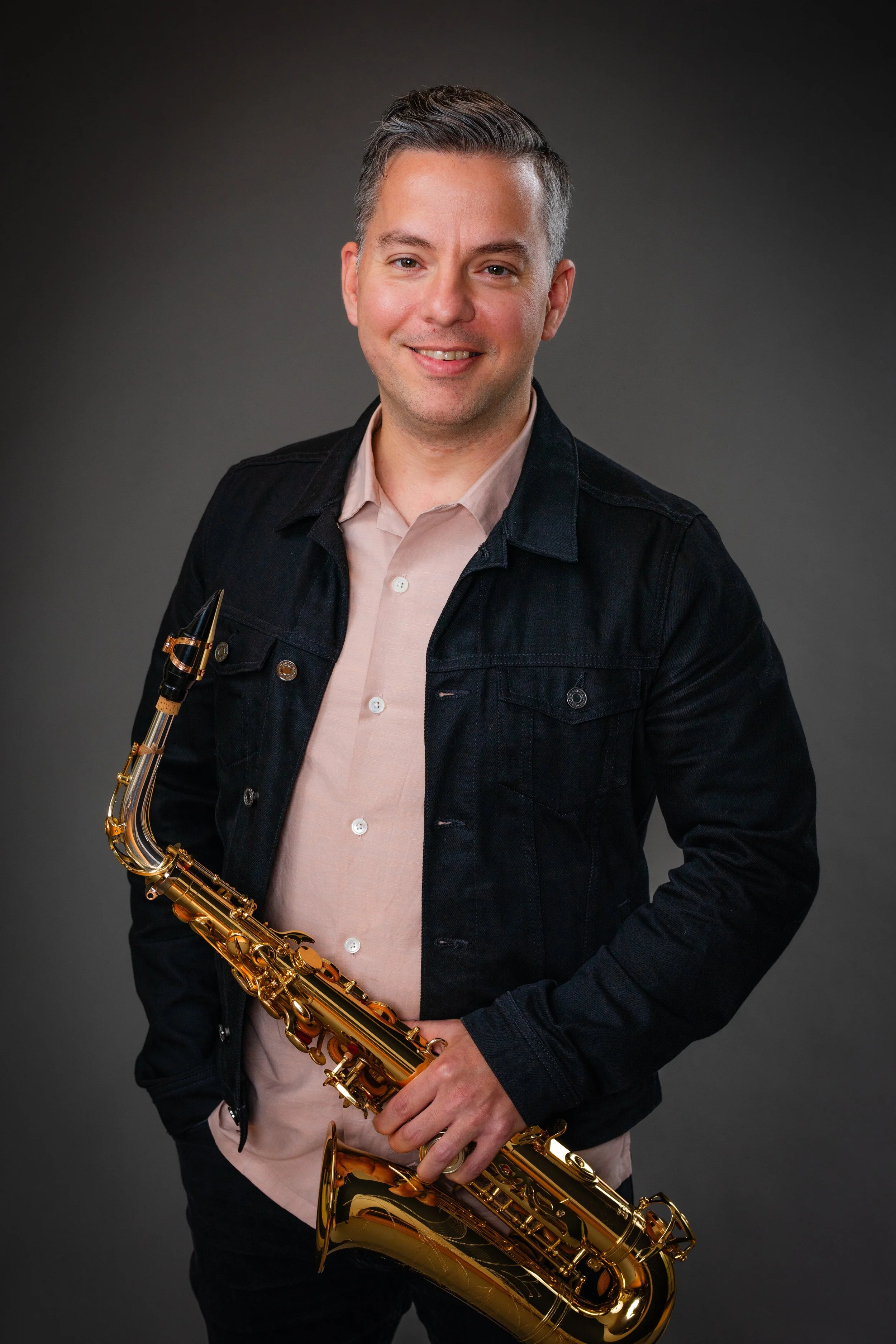 A man with short dark hair and light skin smiling while holding a gold saxophone, wearing a black denim jacket and a light pink button-up shirt, posed against a dark gray background.
