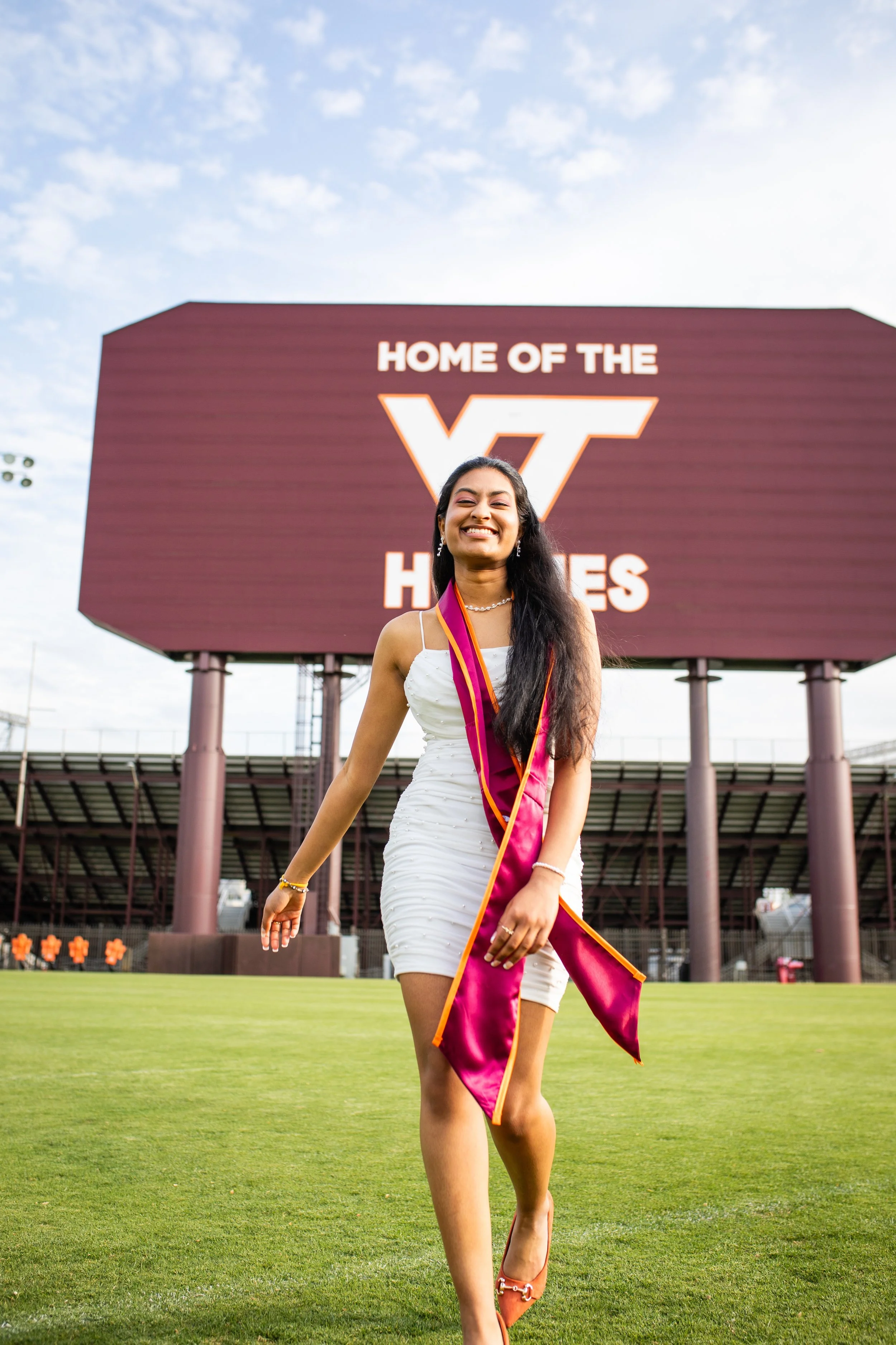 A young woman in a white dress and sash walking on a football field, smiling, with the Virginia Tech Hokies sign in the background.