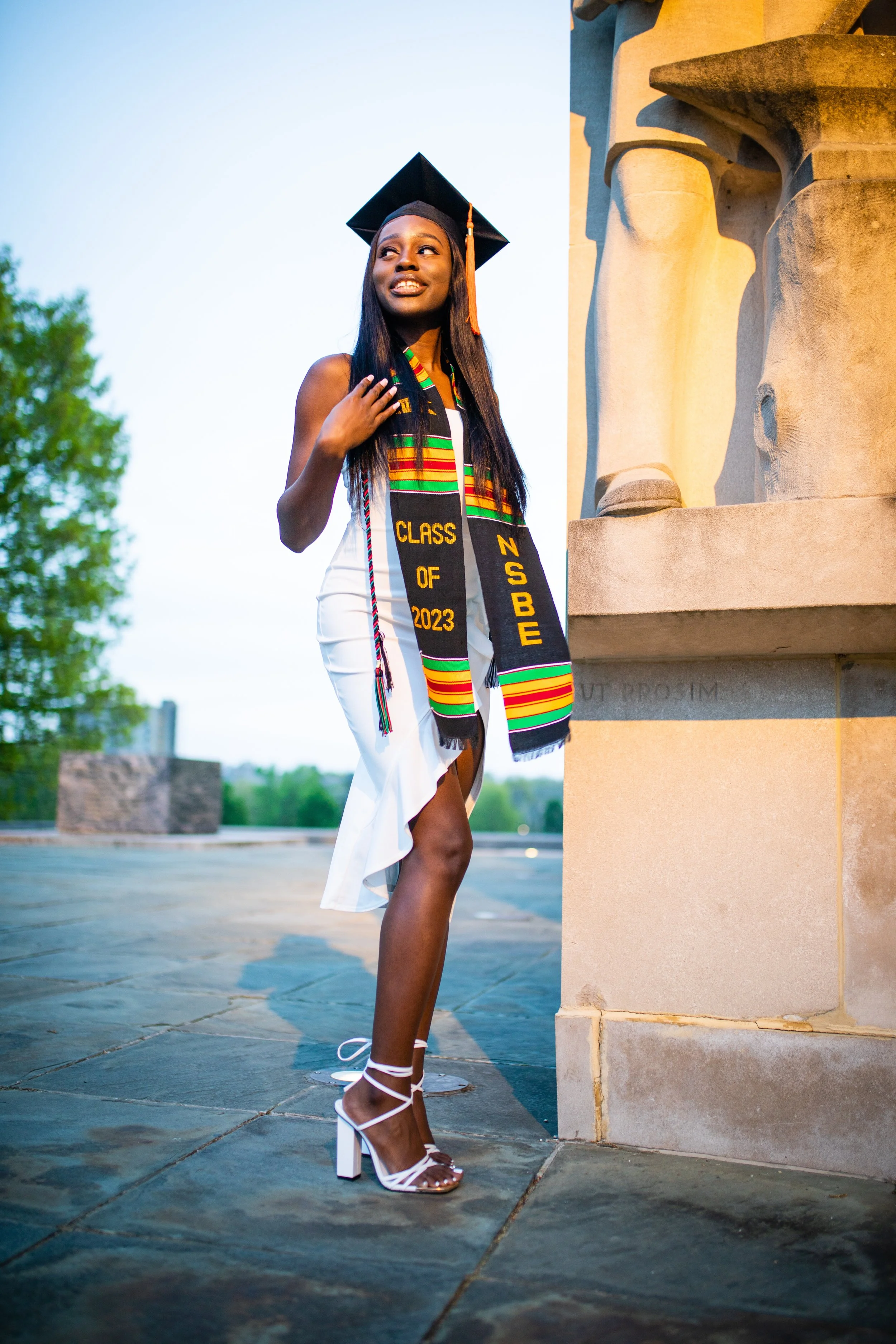 A young woman in a white dress and white high heels performs graduation attire, including a black mortarboard and a multicolored sash that reads 'Class of 2023' and 'NSBE,' standing beside a monument outdoors during the evening.