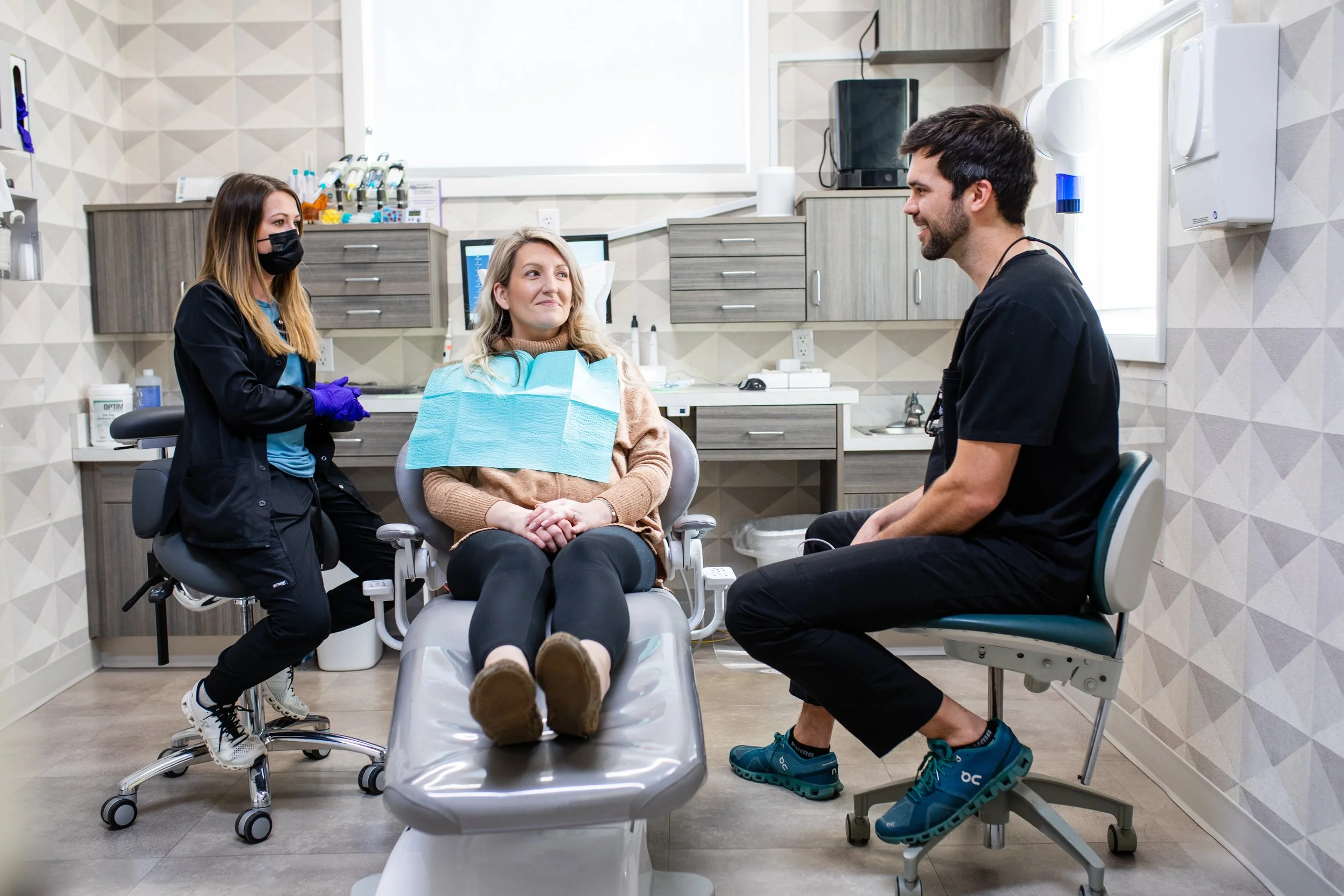 A female patient with a dental bib sits on a dental chair, talking to a male dentist seated on a stool in a dental clinic. A female dental assistant with a face mask and gloves stands nearby, watching the interaction.