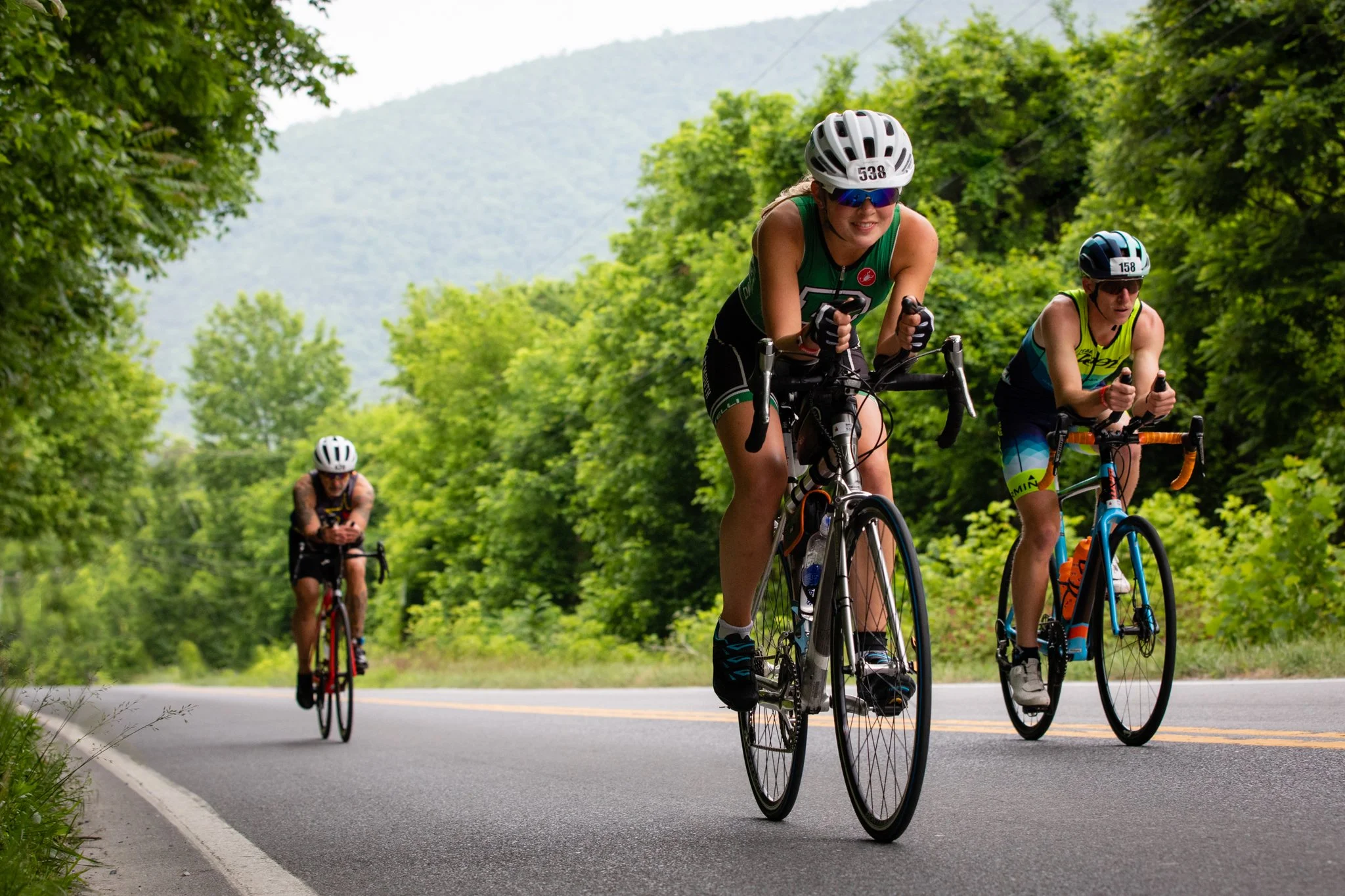 Three cyclists riding on a paved road surrounded by green trees and mountains in the background during daytime.