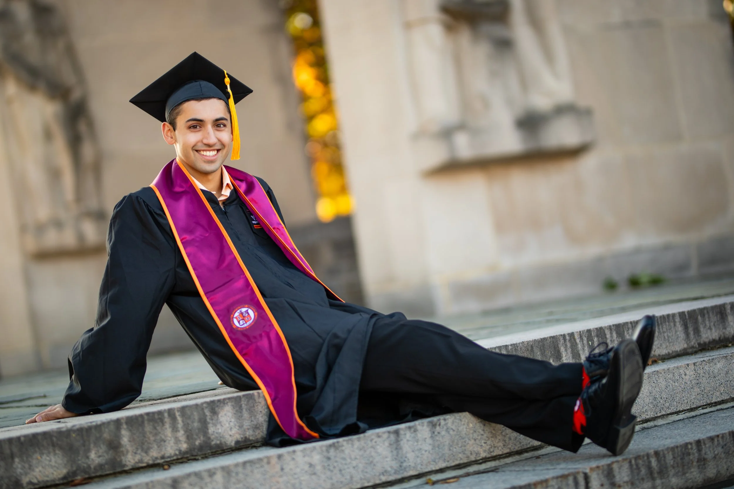 Young man in graduation gown and cap sitting on steps outside, smiling, with a purple stole with an emblem.
