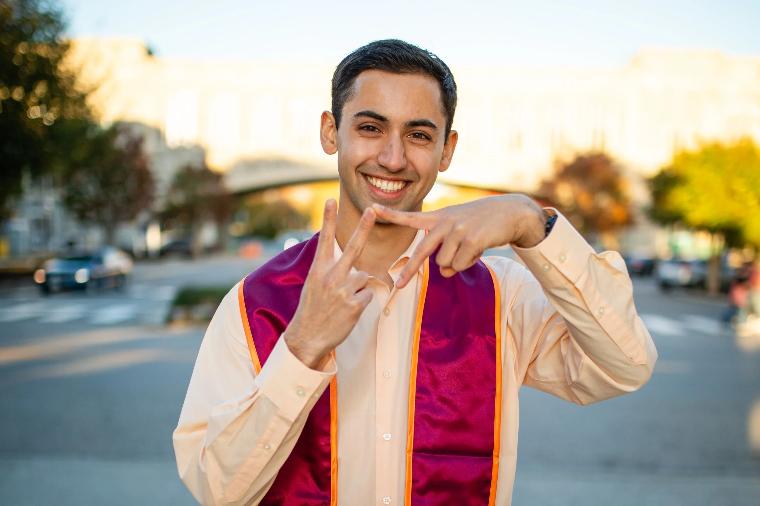 A young man celebrating graduation outdoors, wearing a maroon and orange graduation stole, smiling, making a peace sign with one hand while pointing to himself with the other at sunset.
