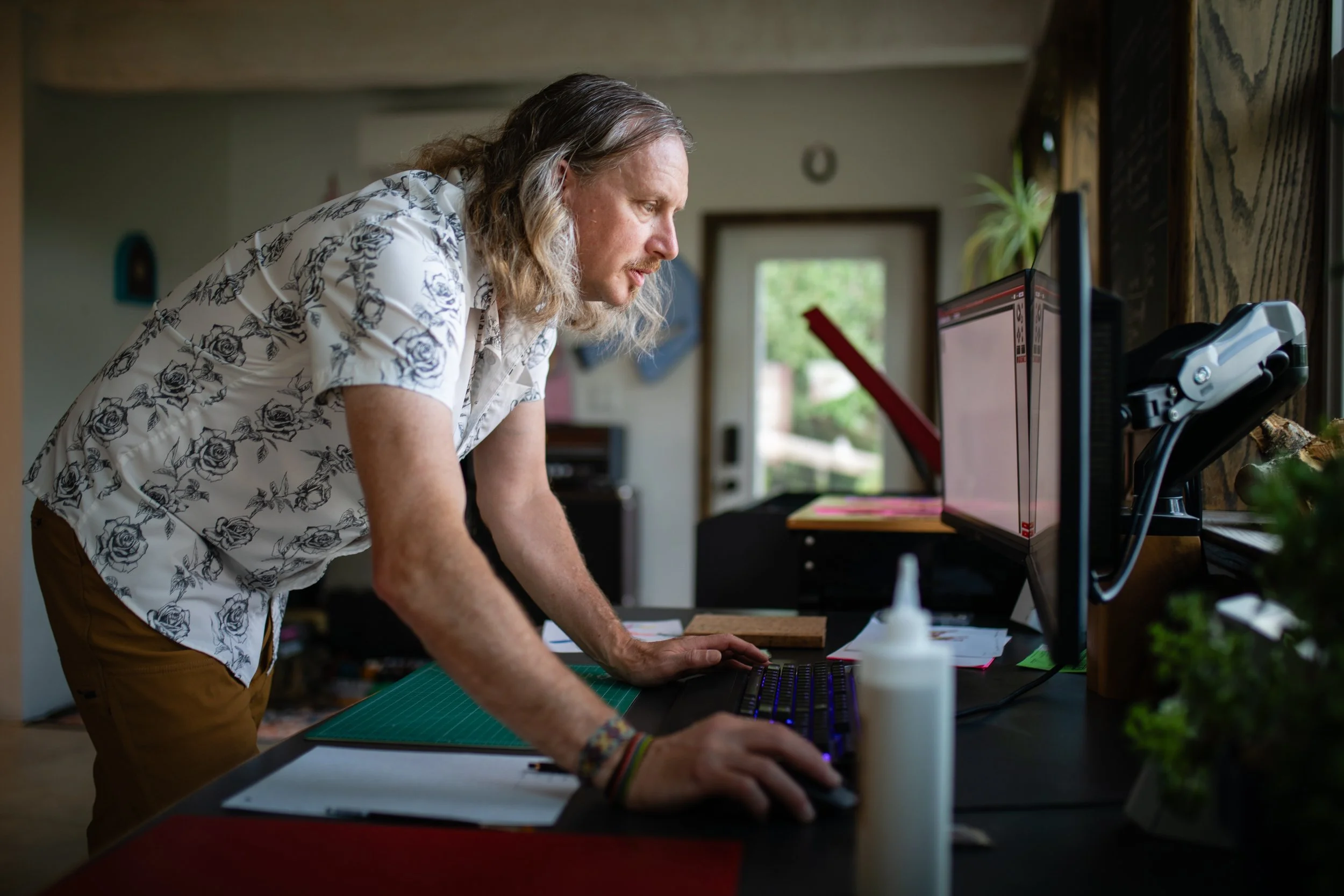 A man with long gray hair leaning over a desk with a computer monitor, keyboard, and various office supplies in a home office.