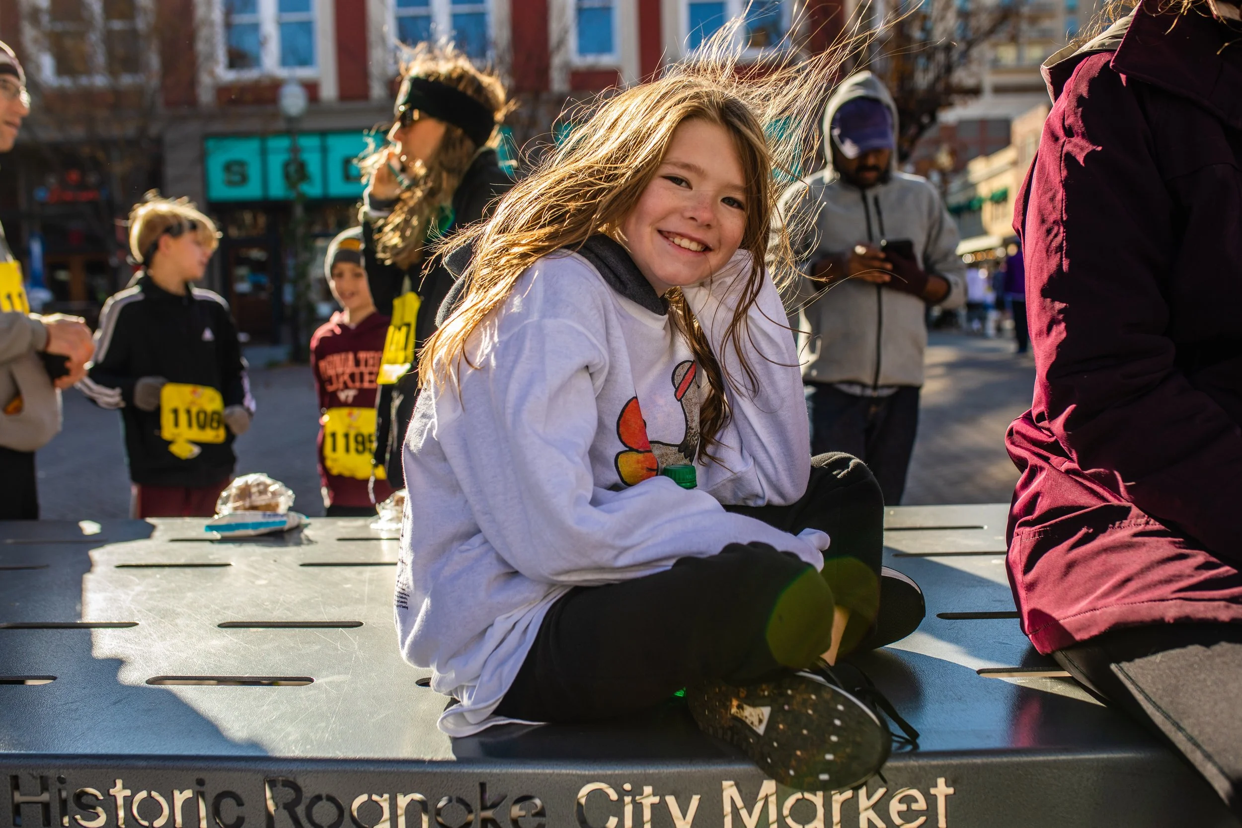 A young girl with long hair, sitting on a table at the Historic Ratonke City Market during a daytime outdoor running event, smiling at the camera with other runners and spectators in the background.