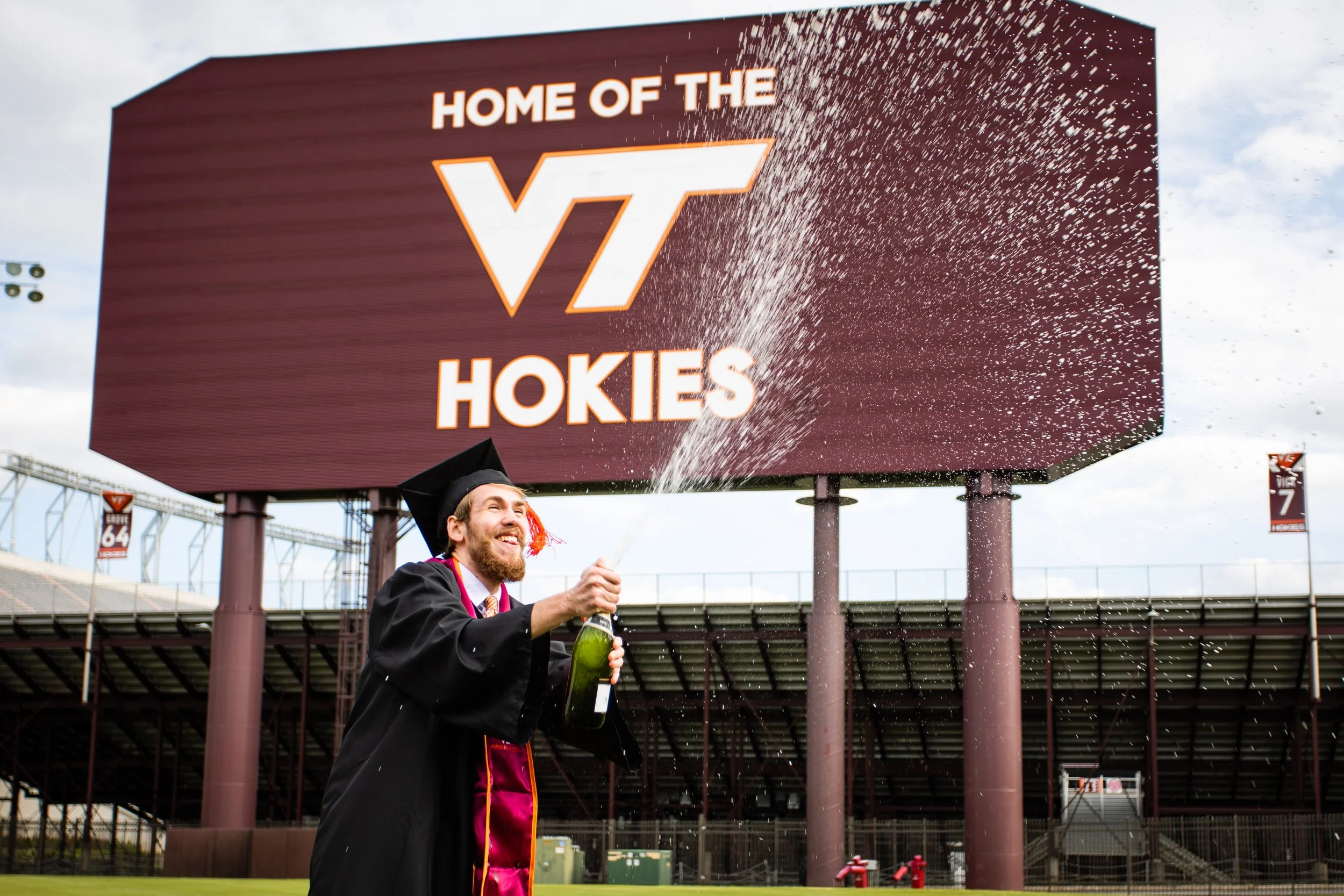 Graduate in cap and gown celebrating with champagne in front of scoreboard that reads 'Home of the VT Hokies' at a football stadium.