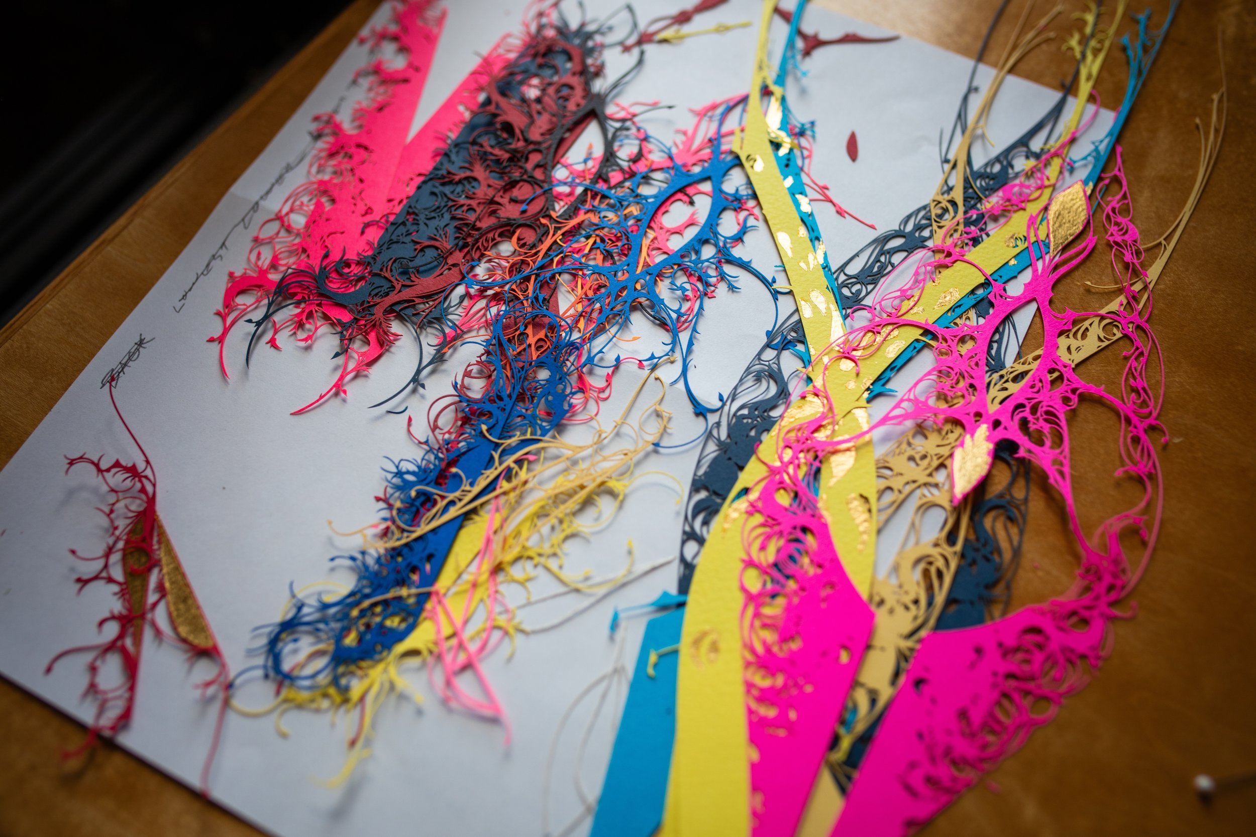 Colorful laser-cut paper strips in pink, black, blue, yellow, and gold, arranged on a white sheet of paper on a wooden table.