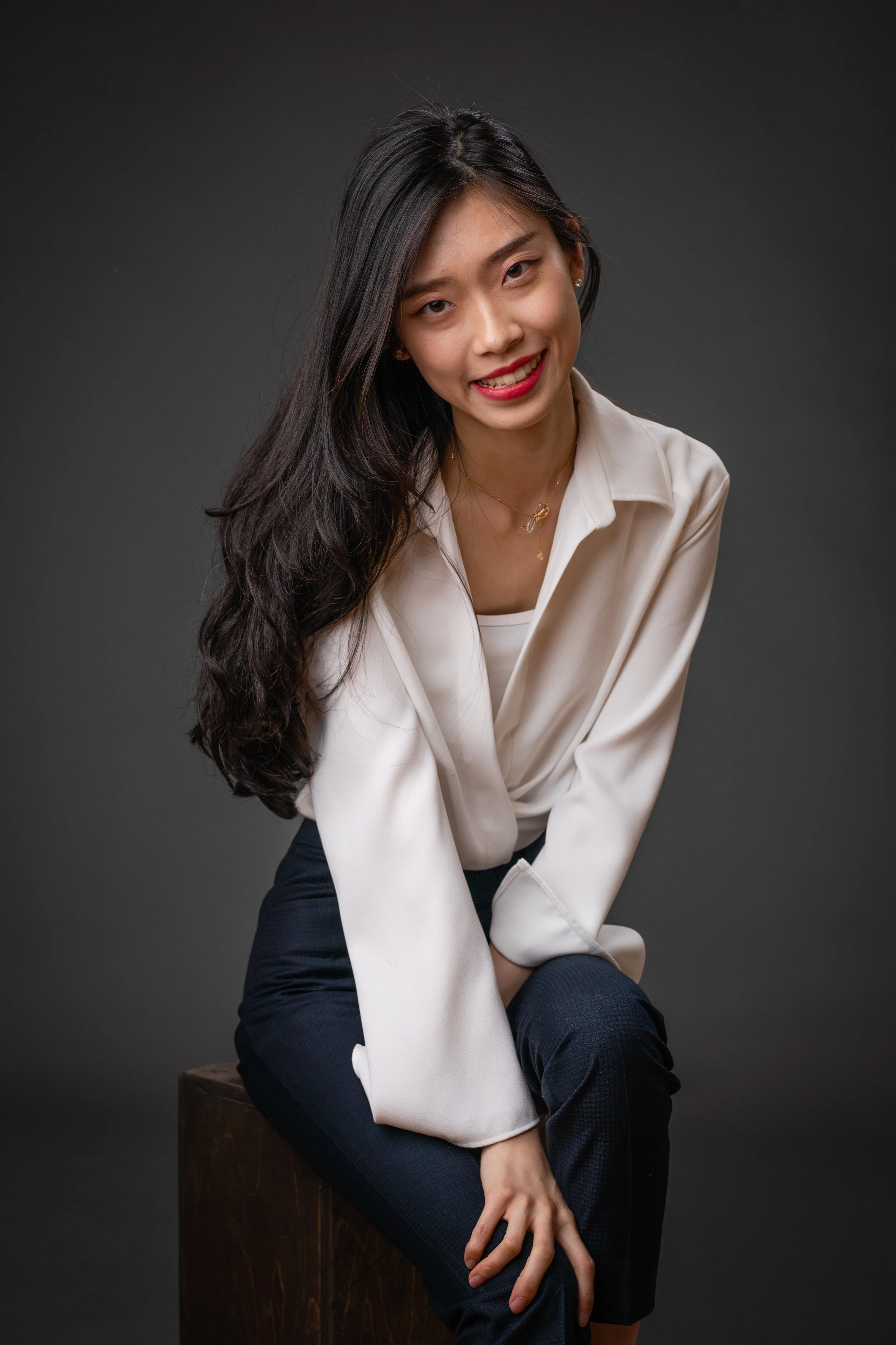 A woman with long dark hair wearing a white blouse and dark pants, sitting on a wooden block against a dark background, smiling at the camera.