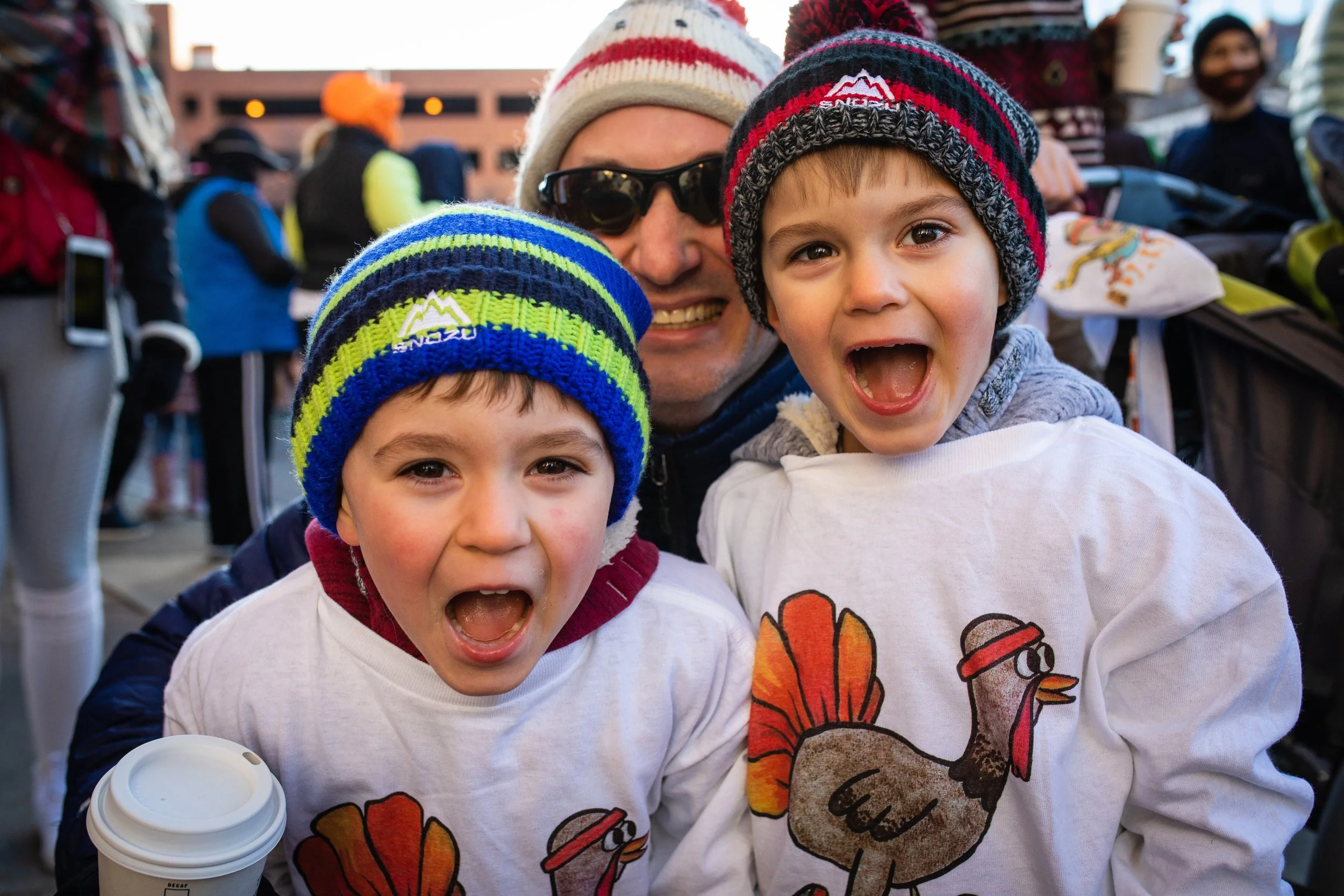 A smiling man with sunglasses and two children wearing winter hats and turkey-themed sweatshirts, standing in a crowd outdoors during cold weather.