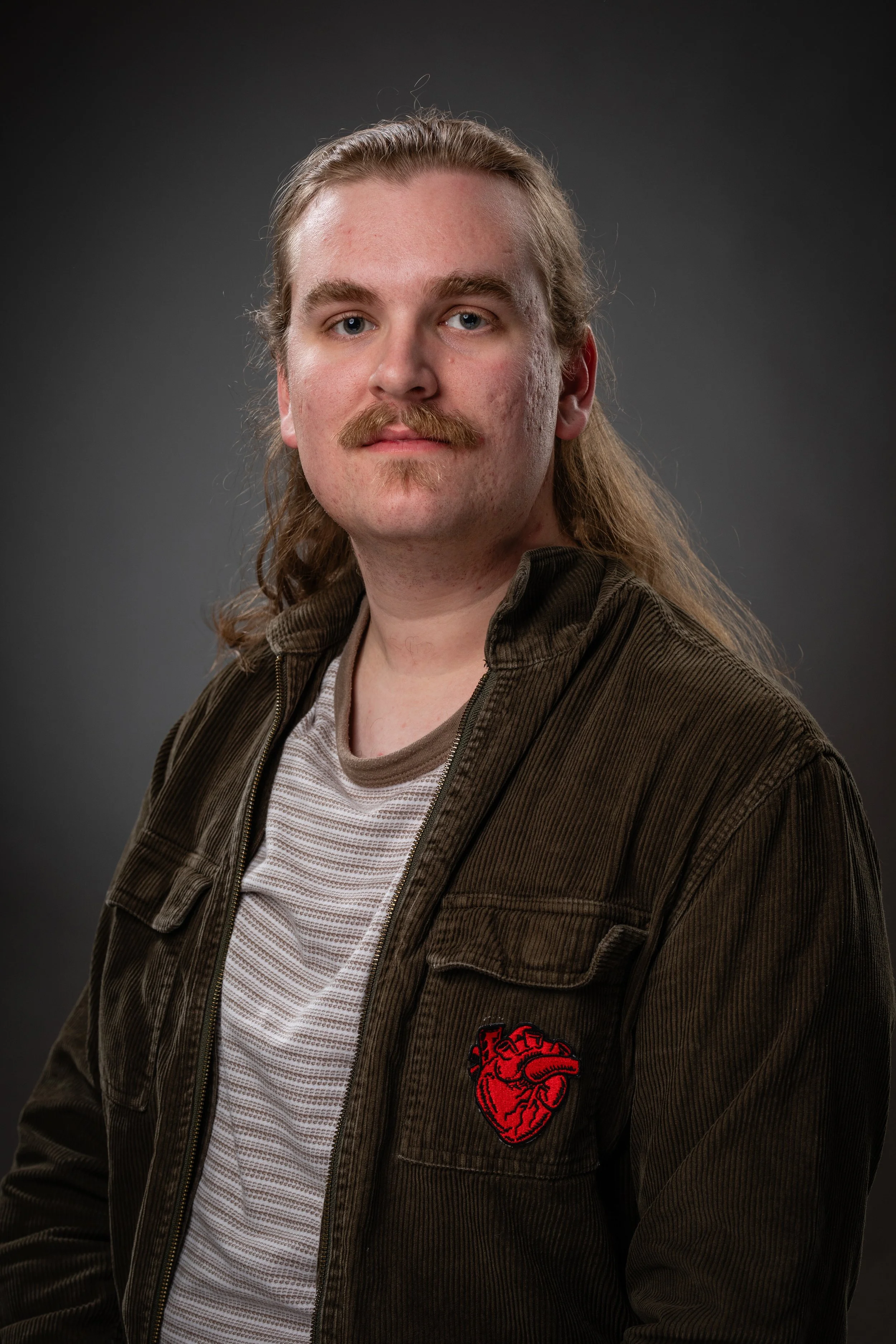Portrait of a man with long hair, mustache, wearing a brown jacket with a red heart emblem, standing against a dark background.