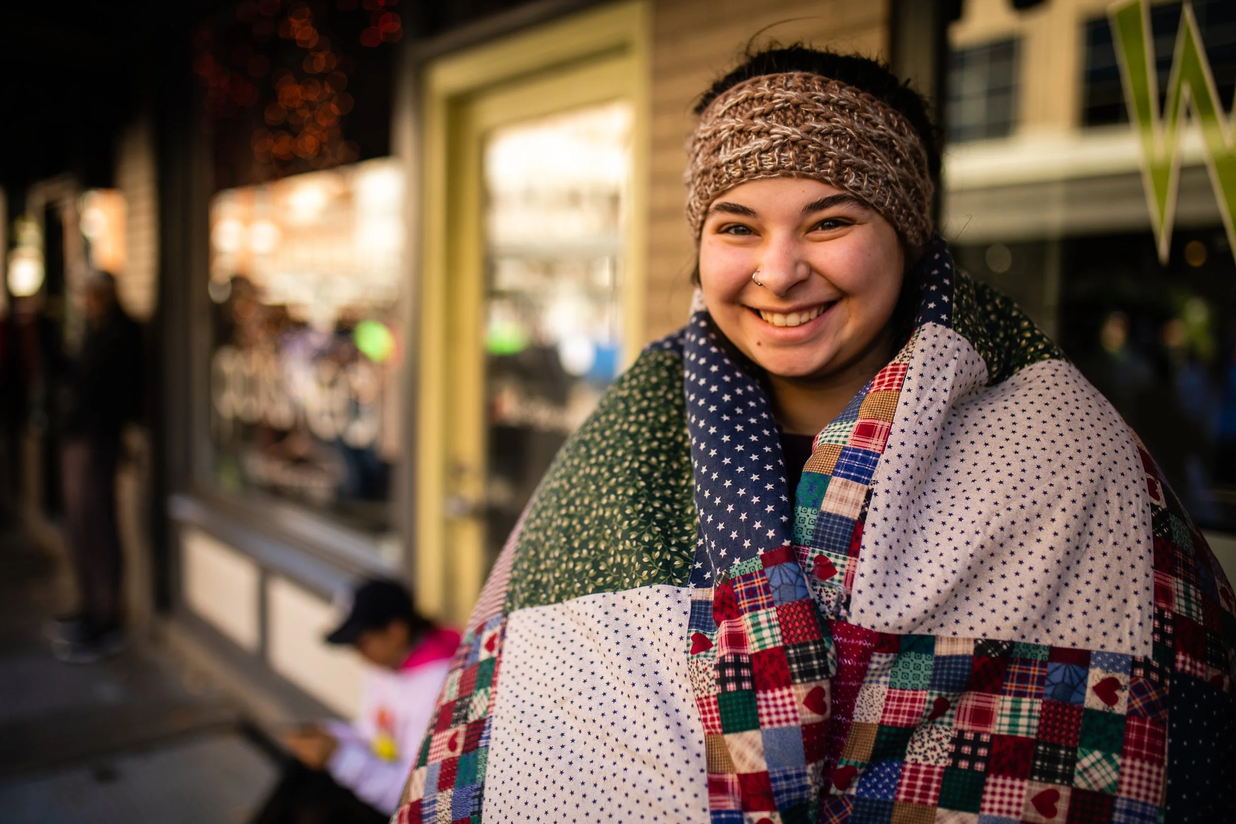 A smiling young woman with a patterned headband and a colorful patchwork quilt draped over her shoulders, standing outdoors during the daytime.