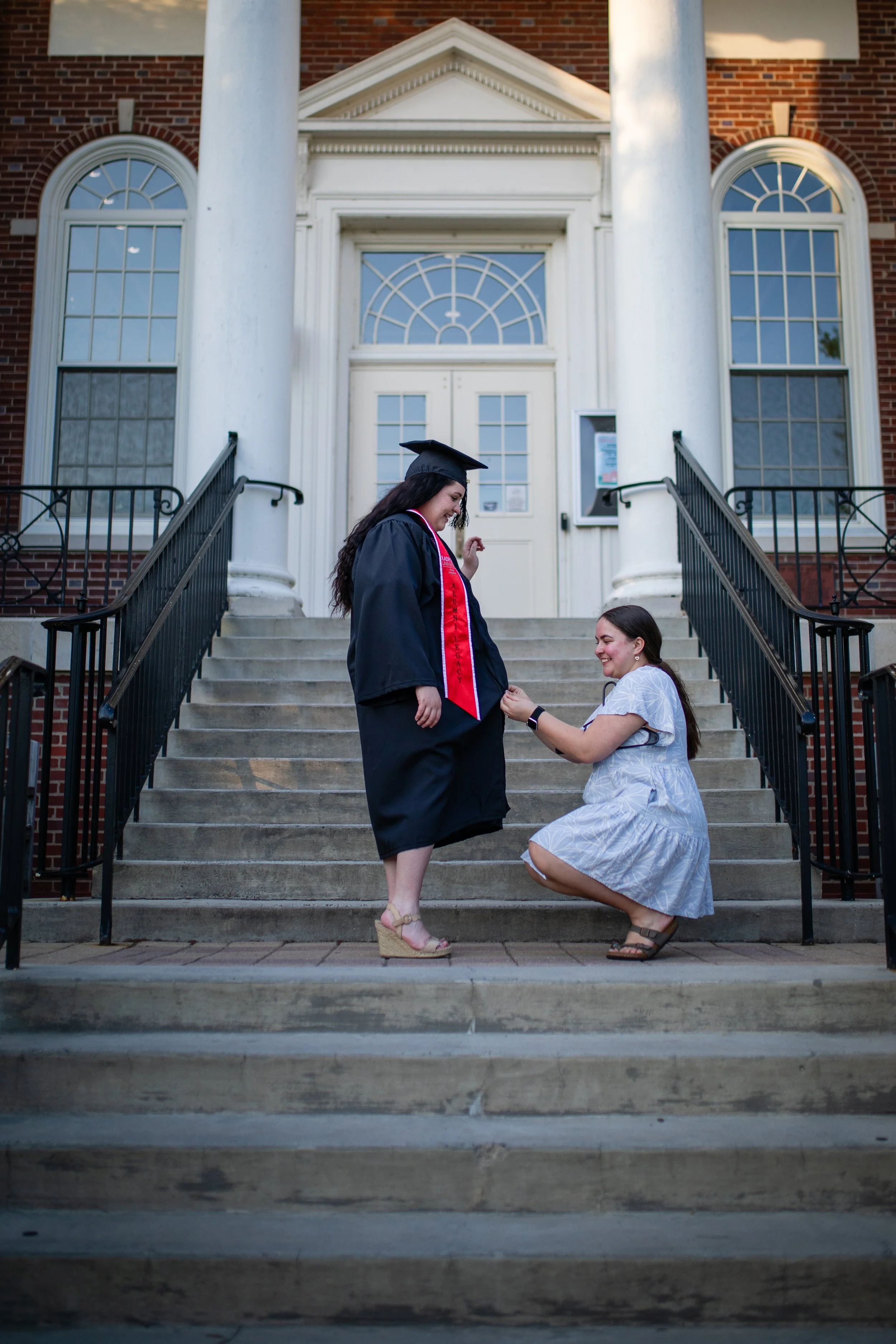 A woman in graduation attire receiving a ring from another woman on the steps of a brick university building.