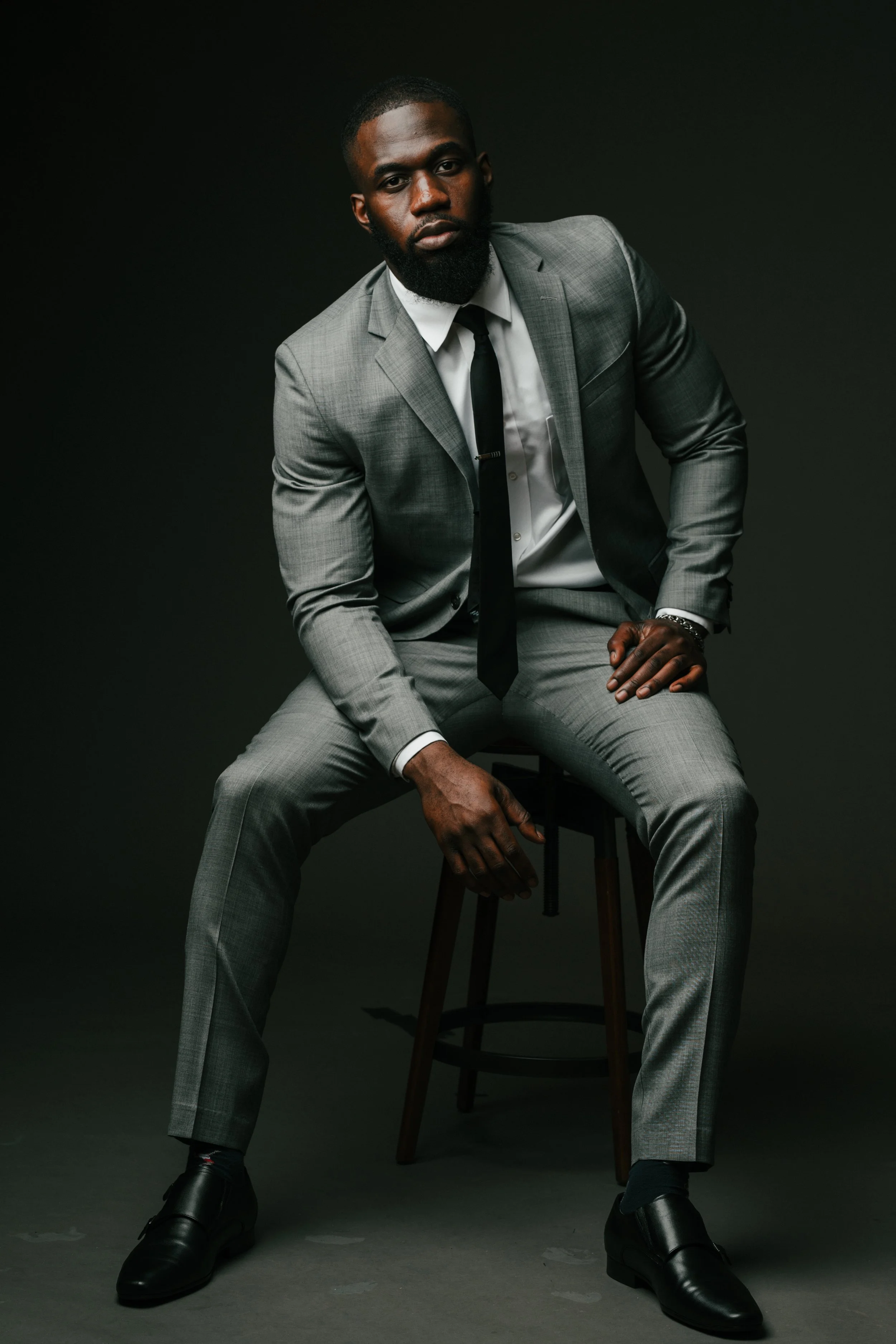 A man in a gray suit, white shirt, and black tie sitting on a stool against a dark background.