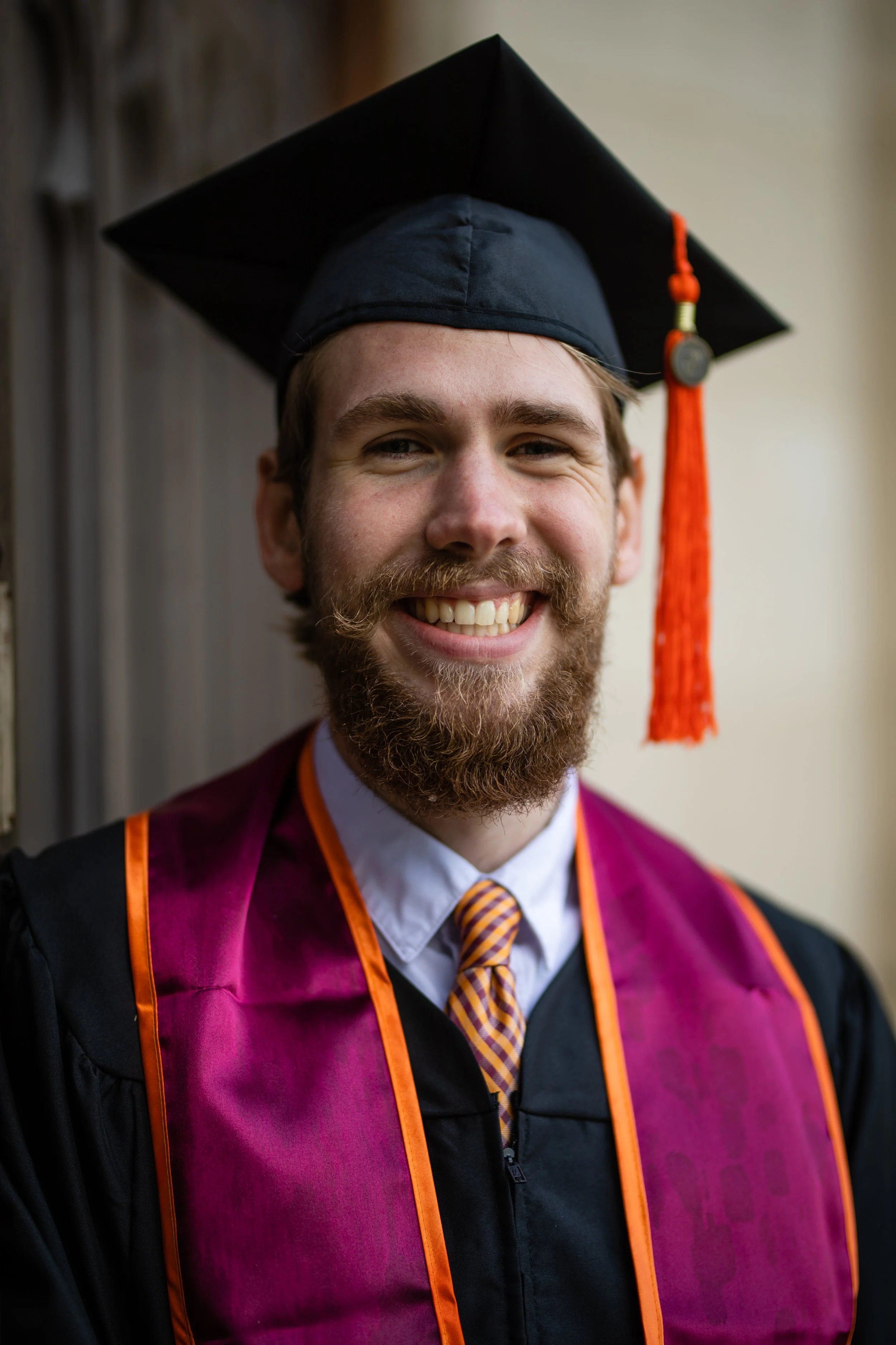 A young man wearing graduation cap and gown, smiling, with a purple and orange stole and a striped tie.