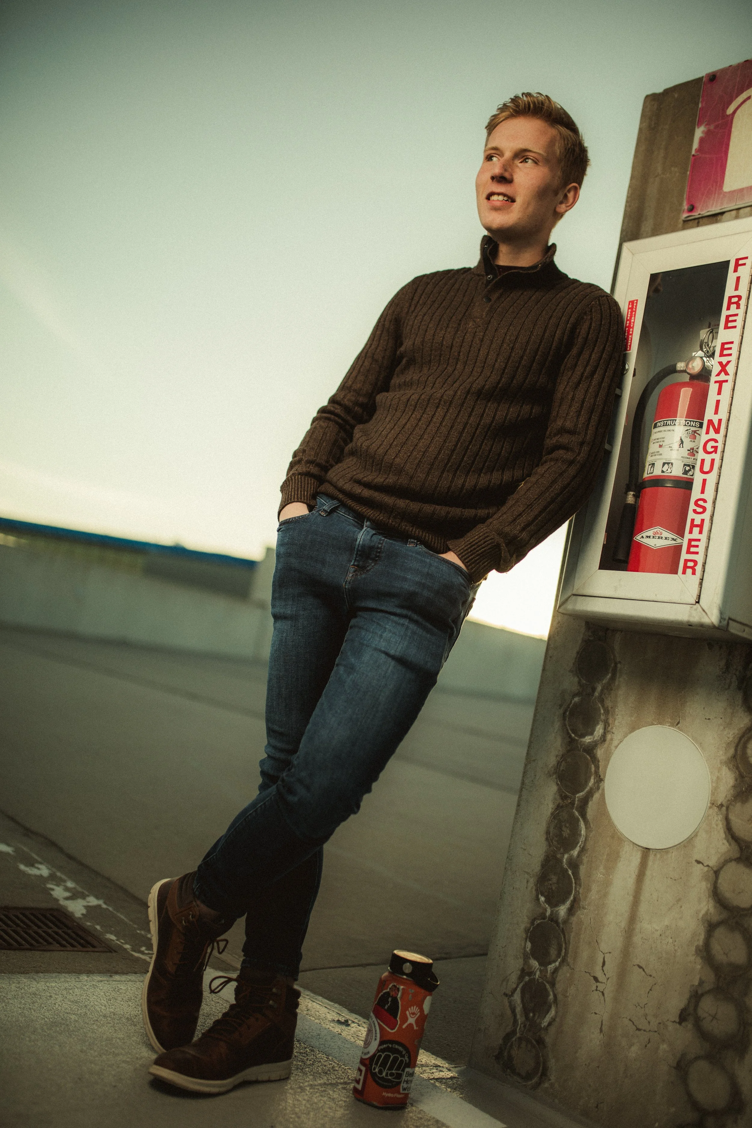 Young man leaning against a fire extinguisher cabinet, wearing a brown sweater, jeans, and brown boots, with a coffee cup on the ground.