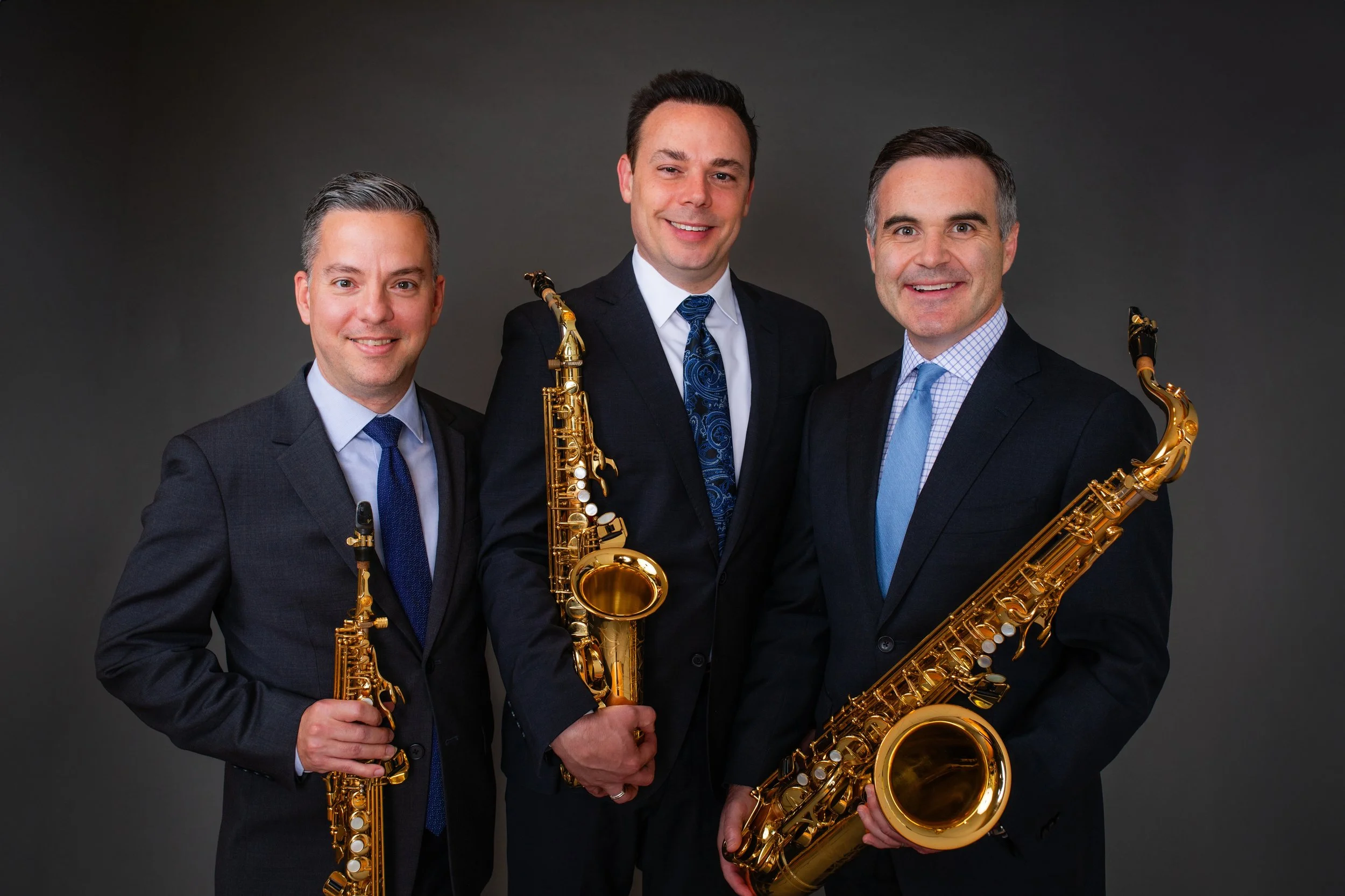 Three men in suits holding saxophones standing against a dark background.