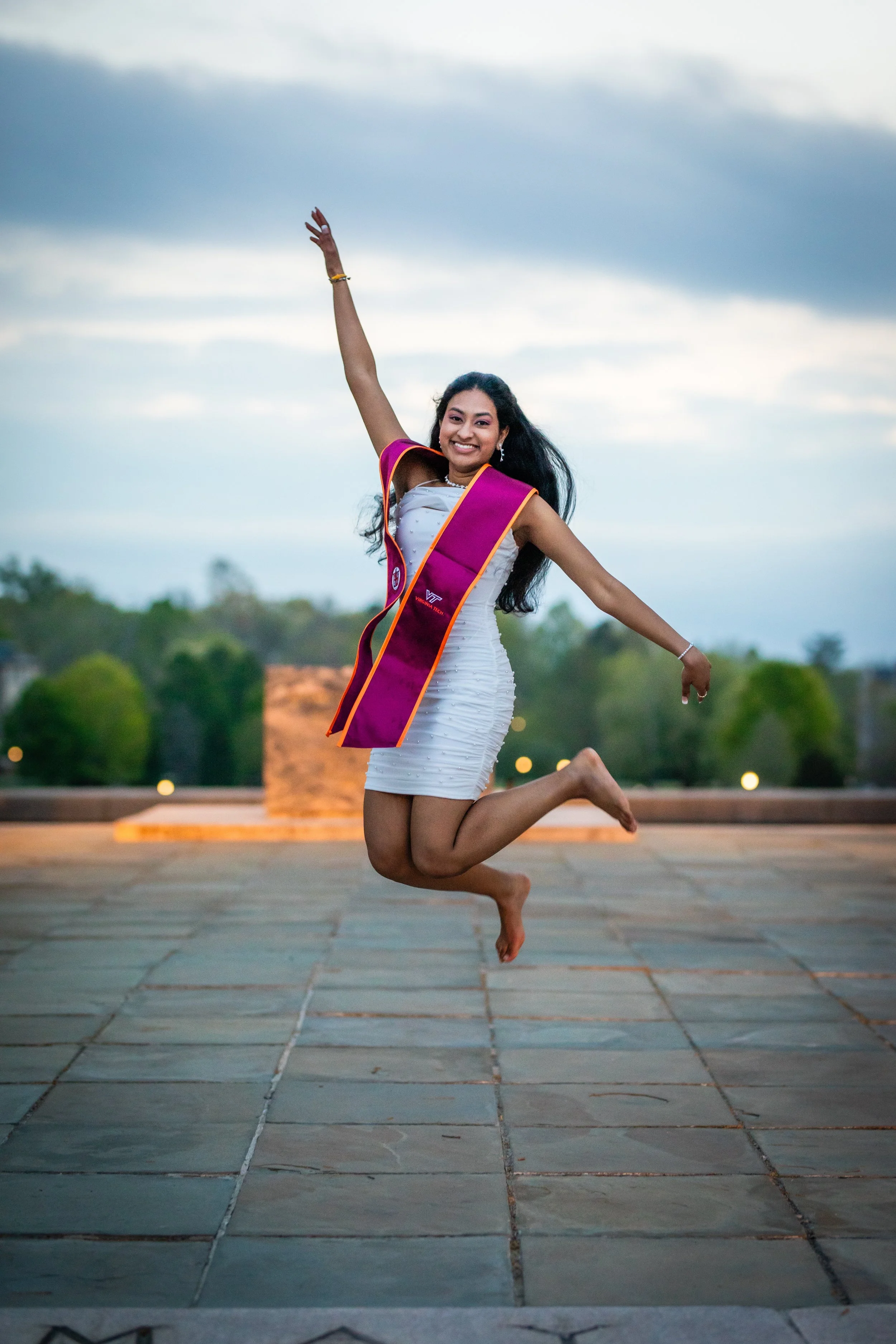 A young woman in a graduation gown and sash is jumping in the air outdoors, smiling with her arms raised and knees bent.