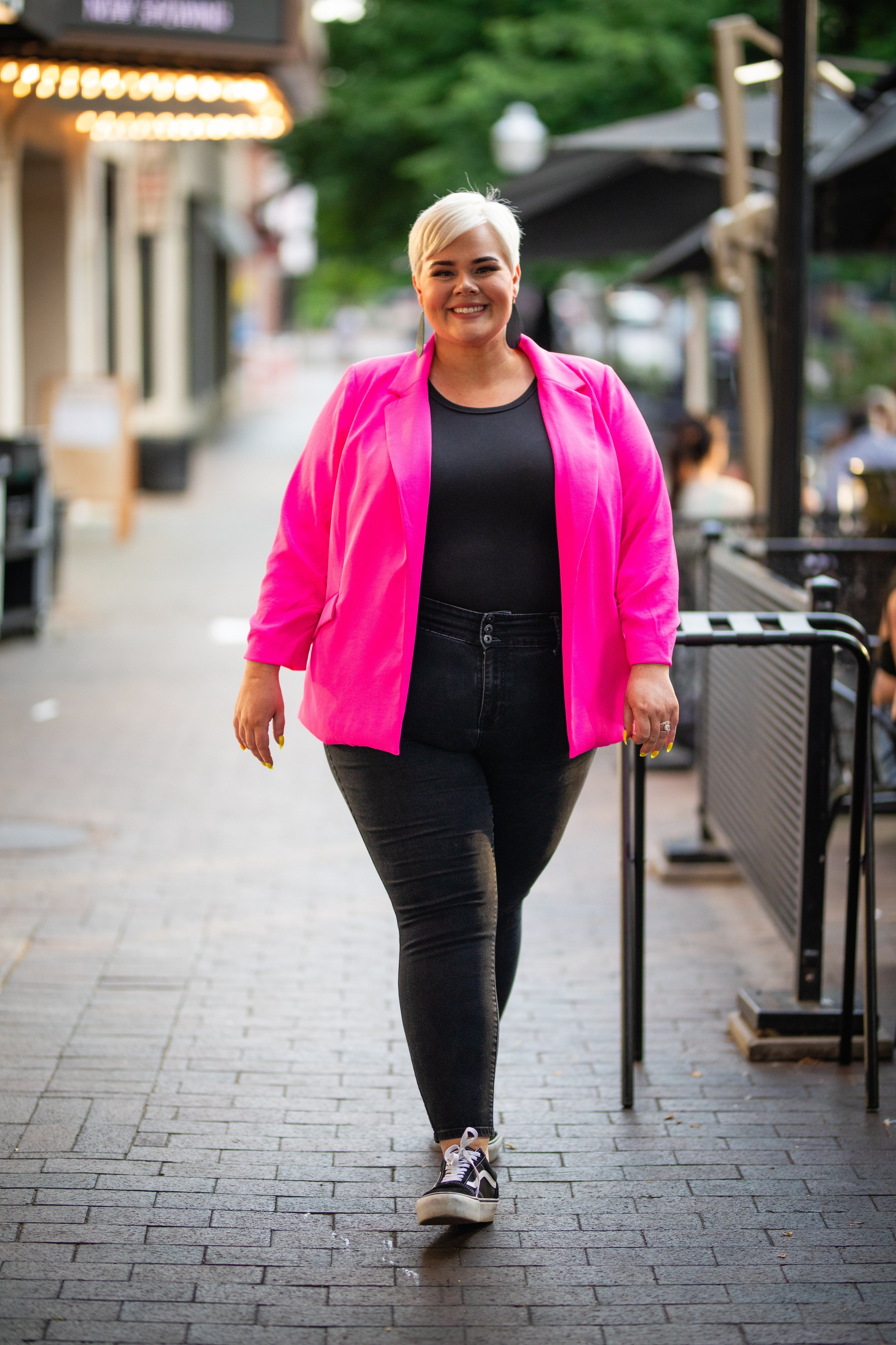 A smiling woman walking down a city sidewalk during the day, wearing a bright pink blazer, black t-shirt, black jeans, and sneakers.