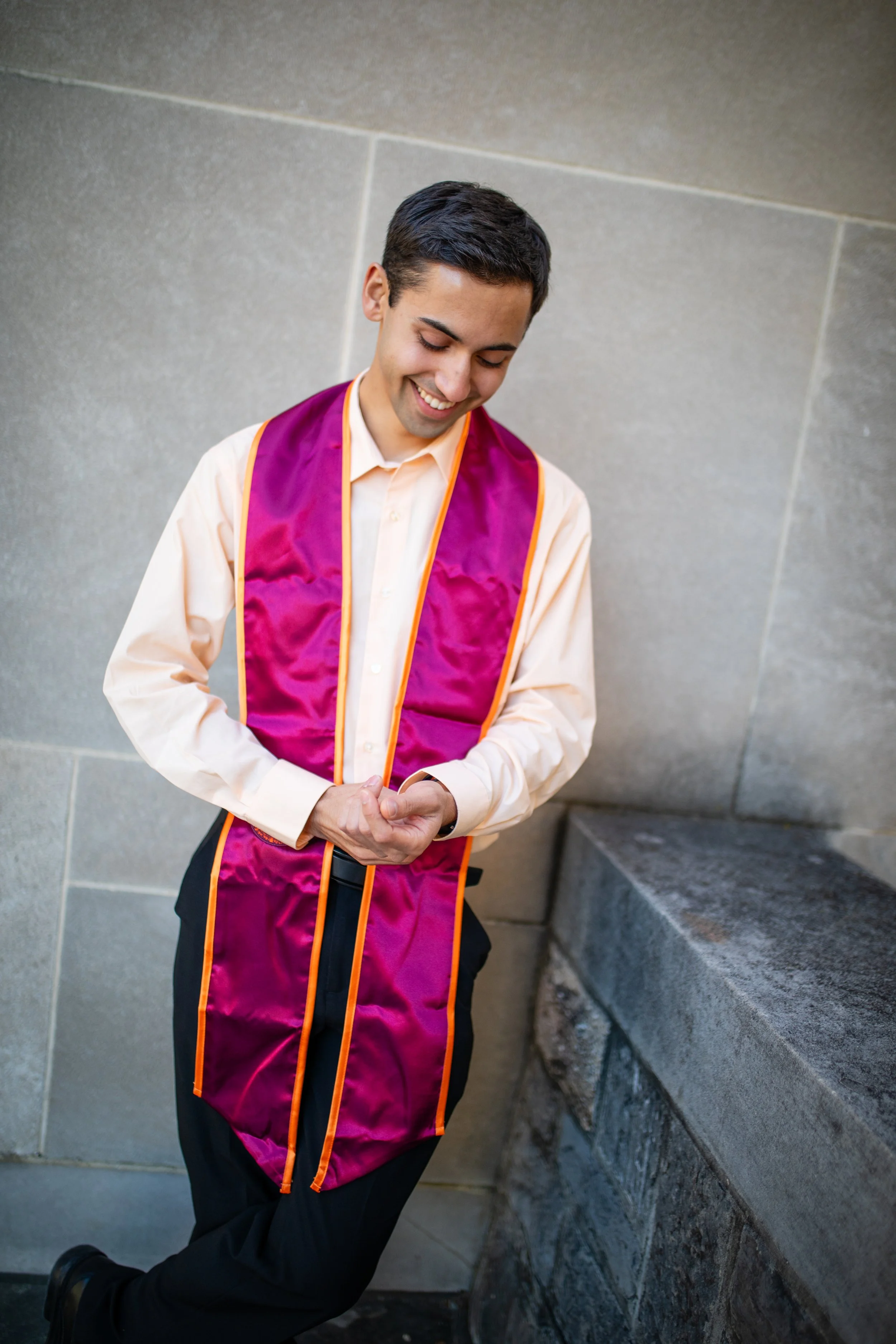 Young man in a white shirt and dark pants wearing a purple and orange graduation stole, standing outside against a stone wall, smiling with eyes closed.