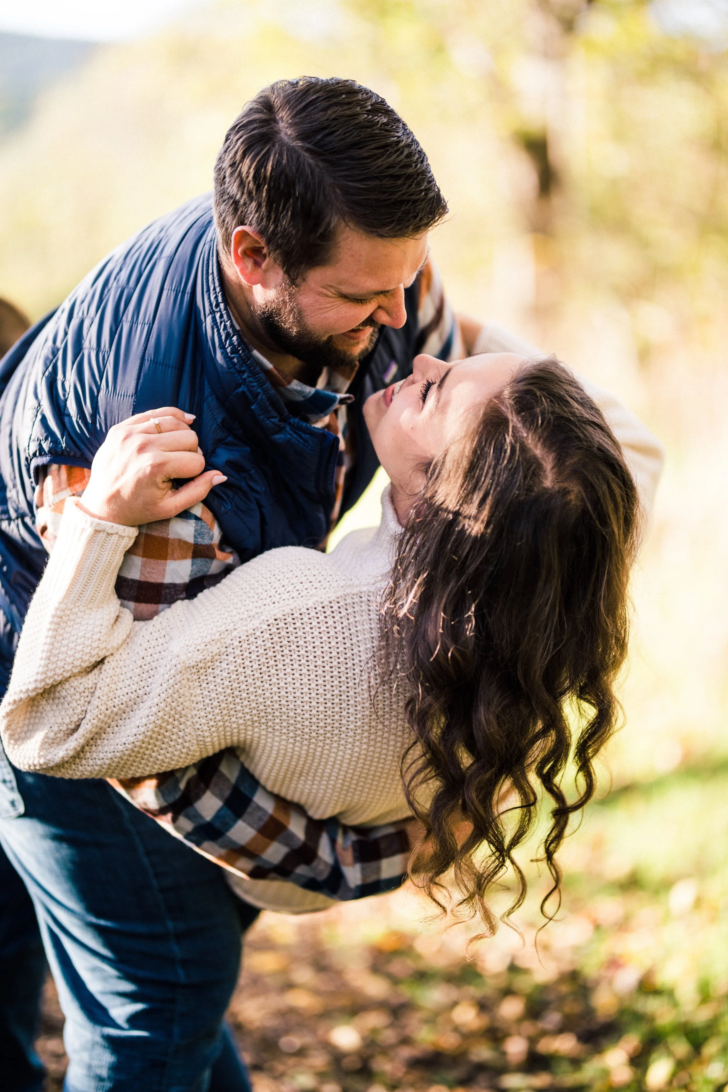 A man and woman outdoors, smiling; the man is holding the woman in a playful dip, both looking at each other with joy.