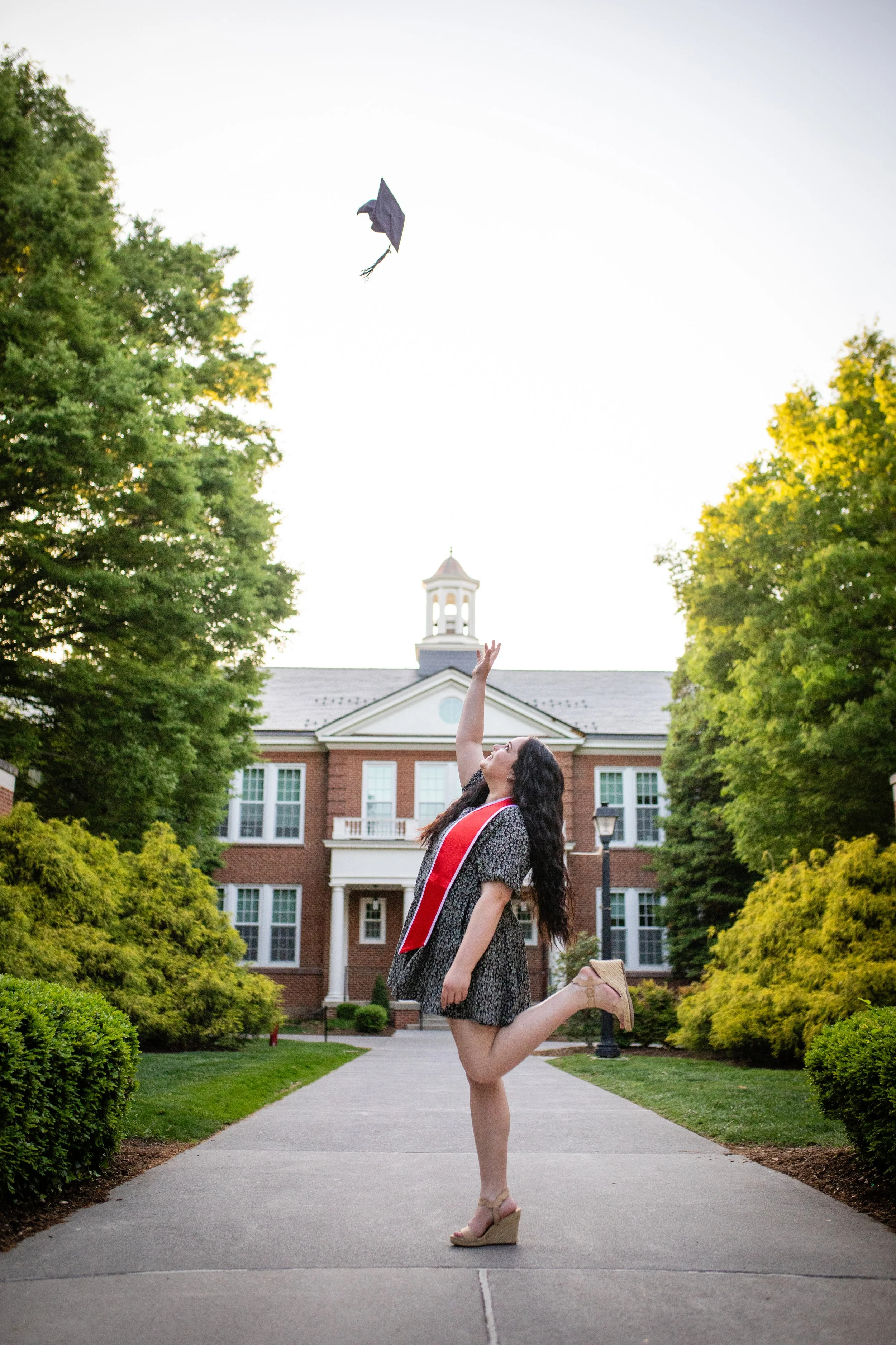 A young woman in a graduation gown and sash throws her cap into the air on campus with greenery and a brick building in the background.