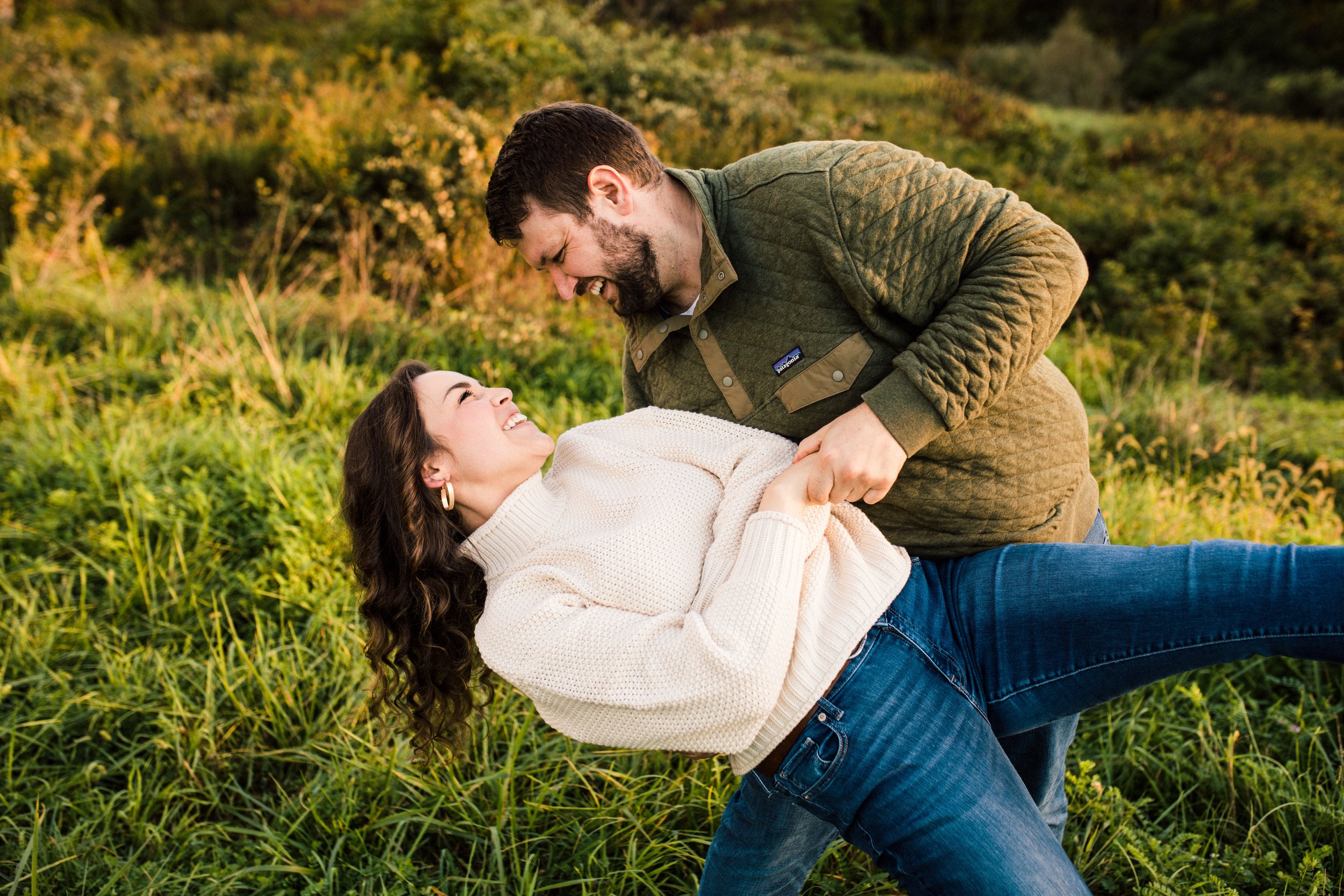 A man and woman are playing and laughing in a grassy outdoor field during sunset, with the man lifting the woman in his arms.