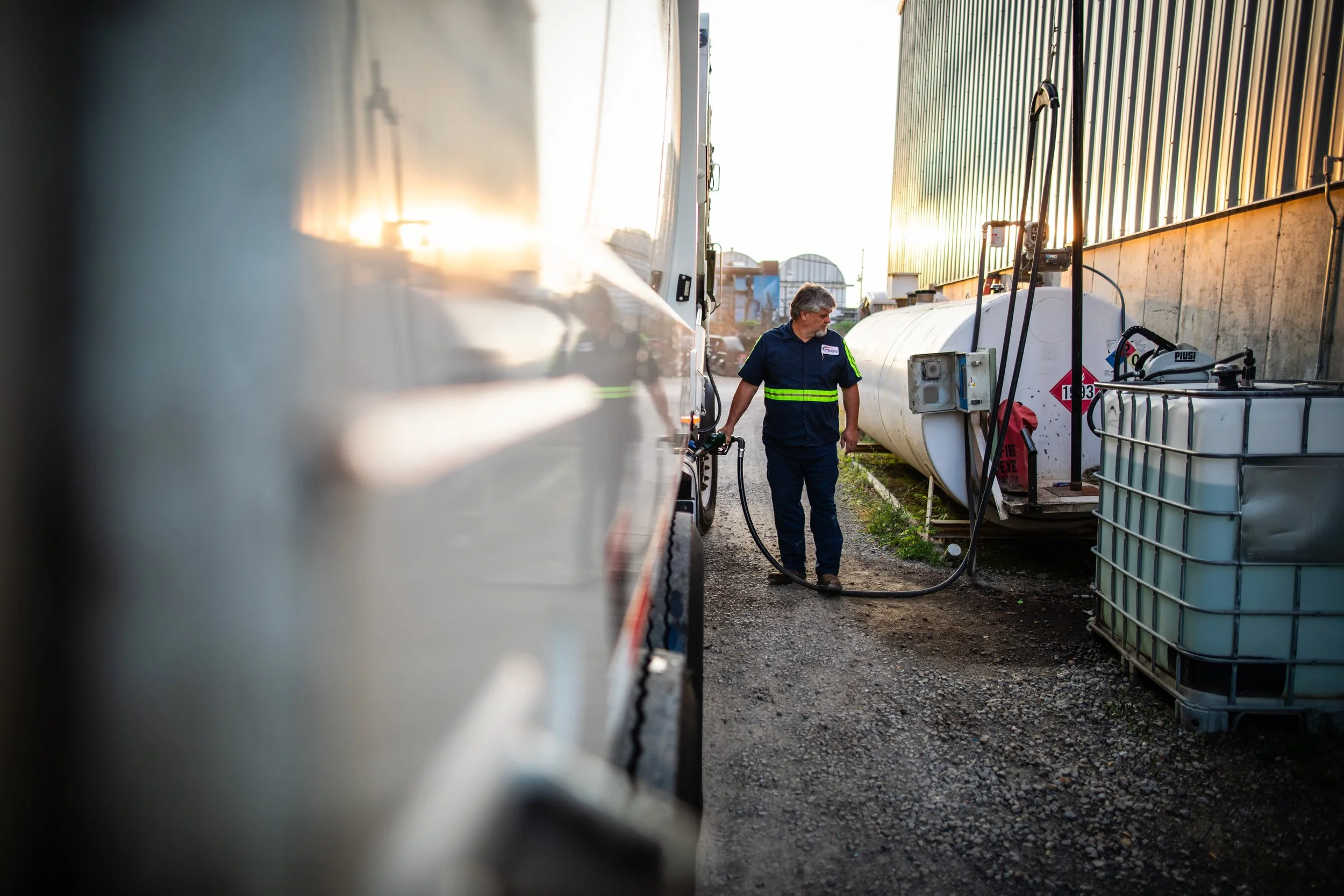 A man in navy uniform with reflective stripes refueling a vehicle from a hose in an industrial area with large tanks and containers nearby.