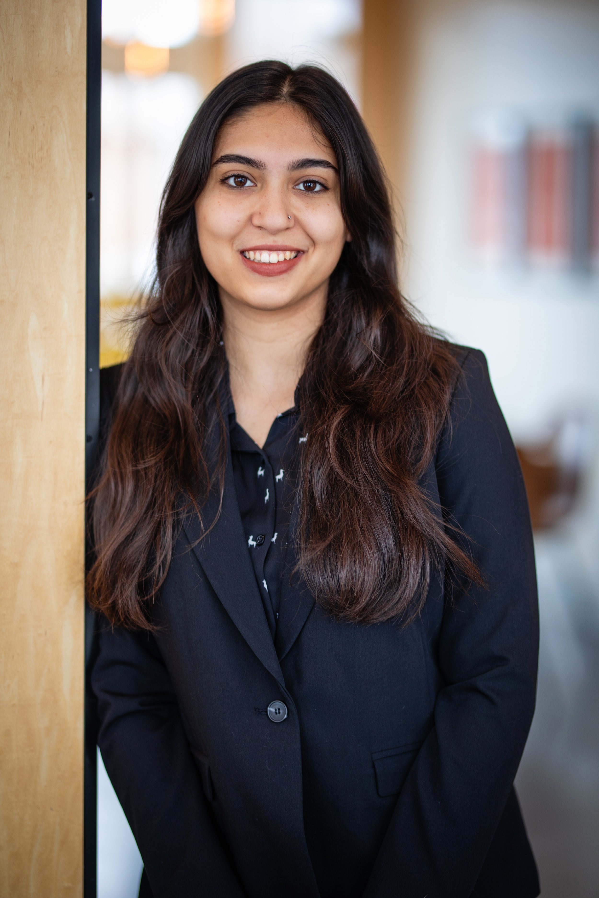 A woman with long dark hair, wearing a black suit, standing indoors near a wooden surface, smiling at the camera.