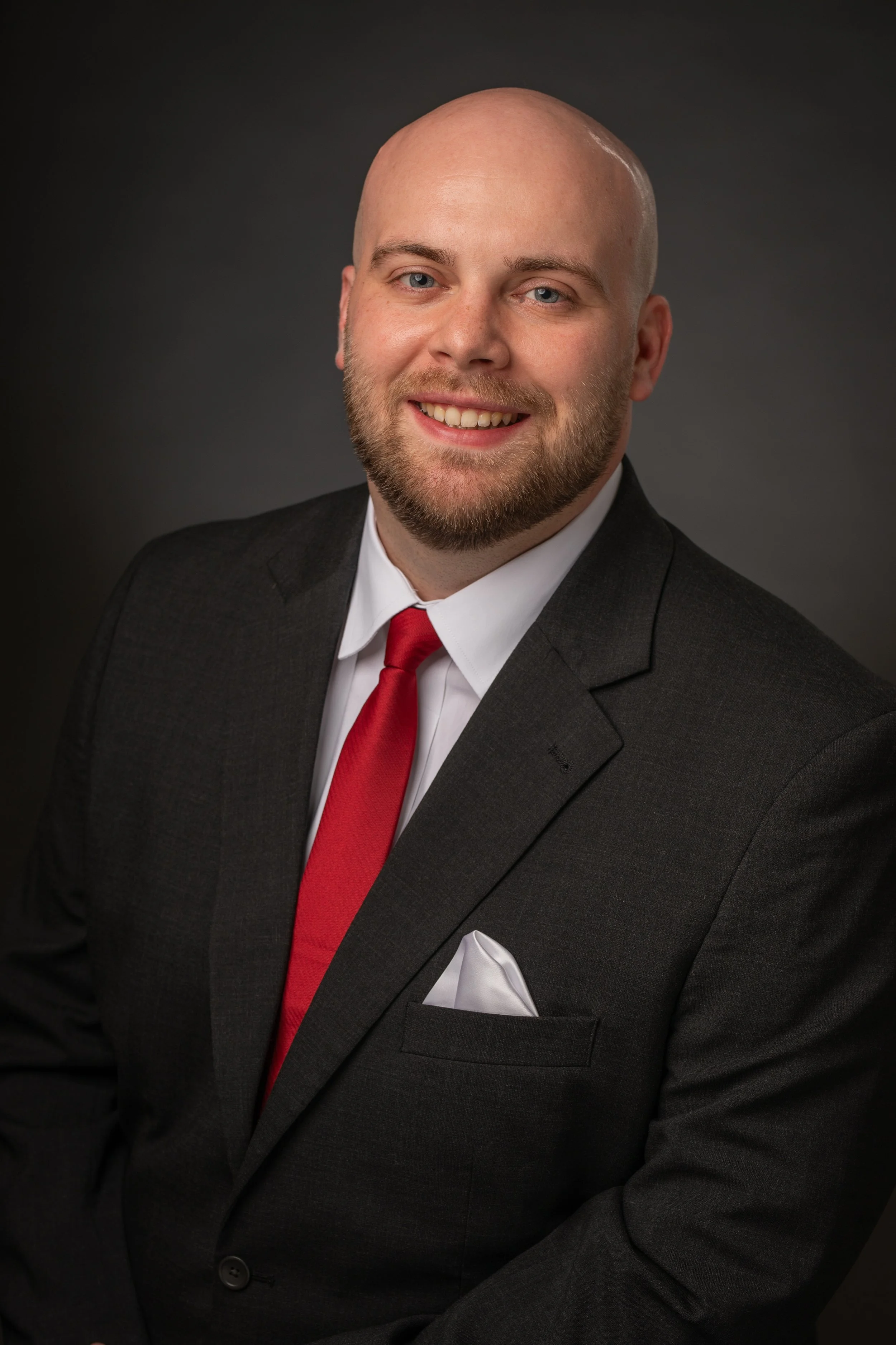 A professional headshot of a bald man with a beard, wearing a dark suit, white shirt, red tie, and white pocket square, smiling against a dark gradient background.