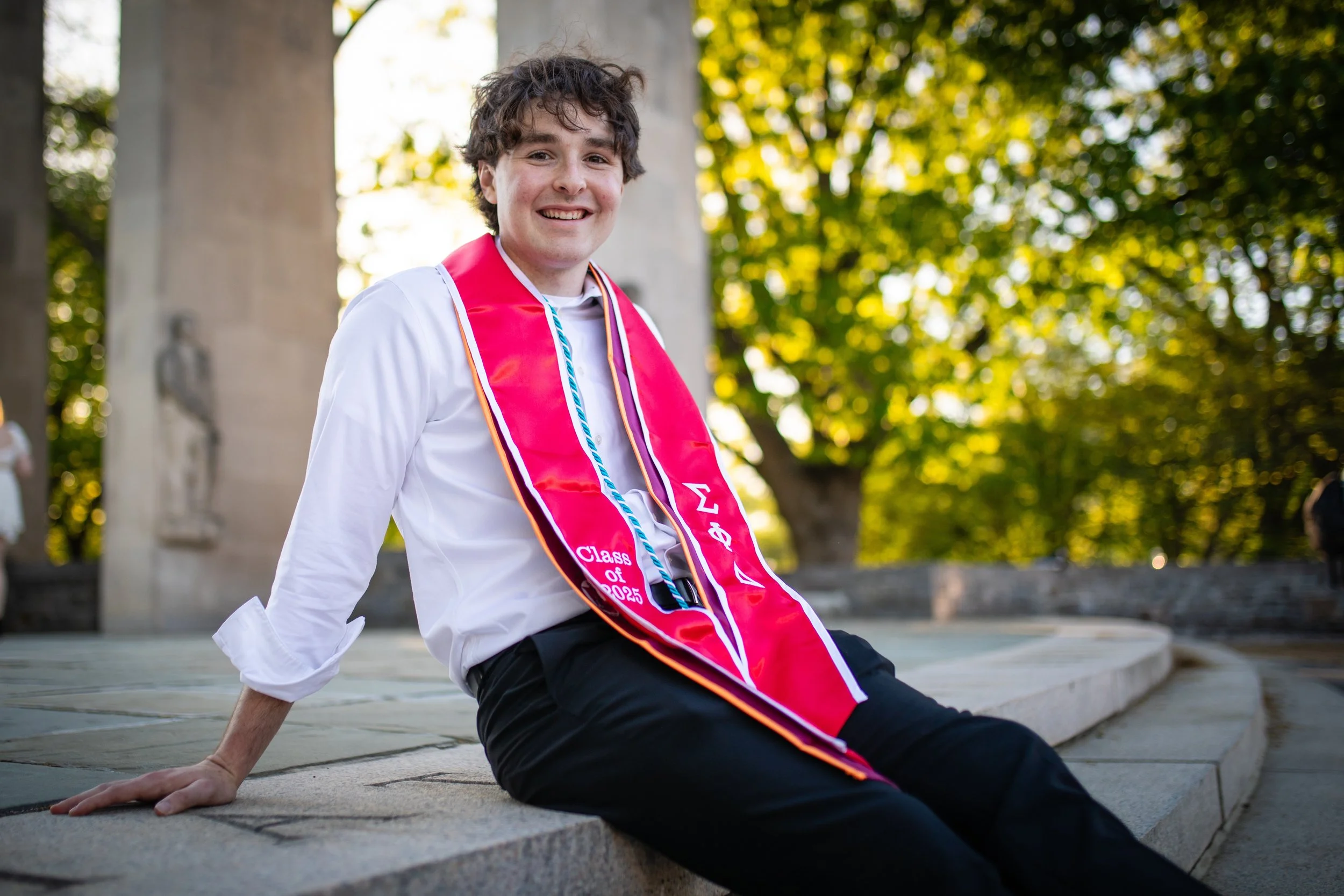 Young man in white shirt and black pants sitting on stone steps outdoors, wearing a pink graduation stole with Greek letters and 'Class of 2023' written on it, smiling at the camera with trees and a monument in the background.