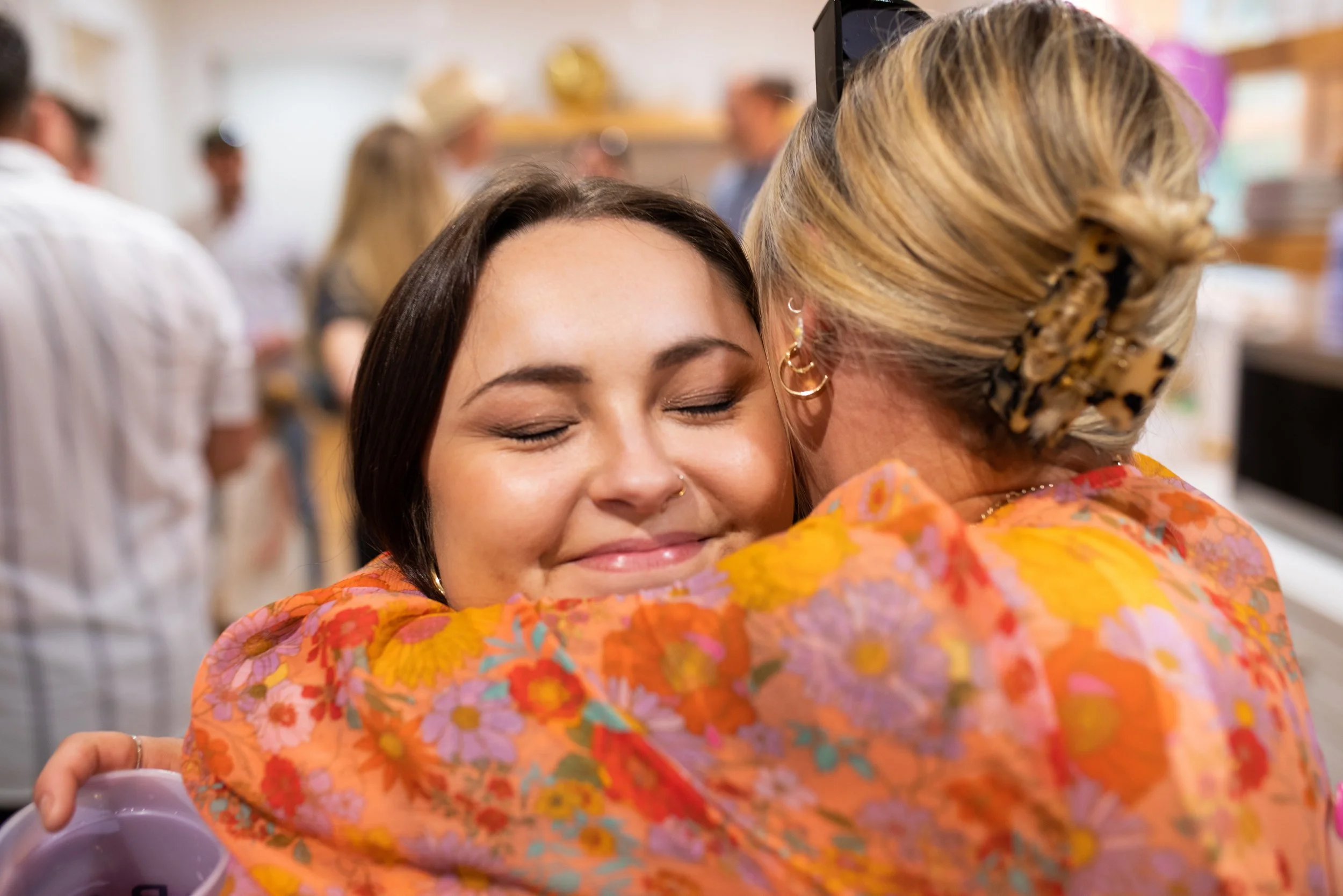 A young woman with dark hair and a nose ring is hugging an older woman with blonde hair in a bun, wearing a colorful floral blouse. The young woman has her eyes closed and a smile on her face. The background shows a busy kitchen or gathering area wit