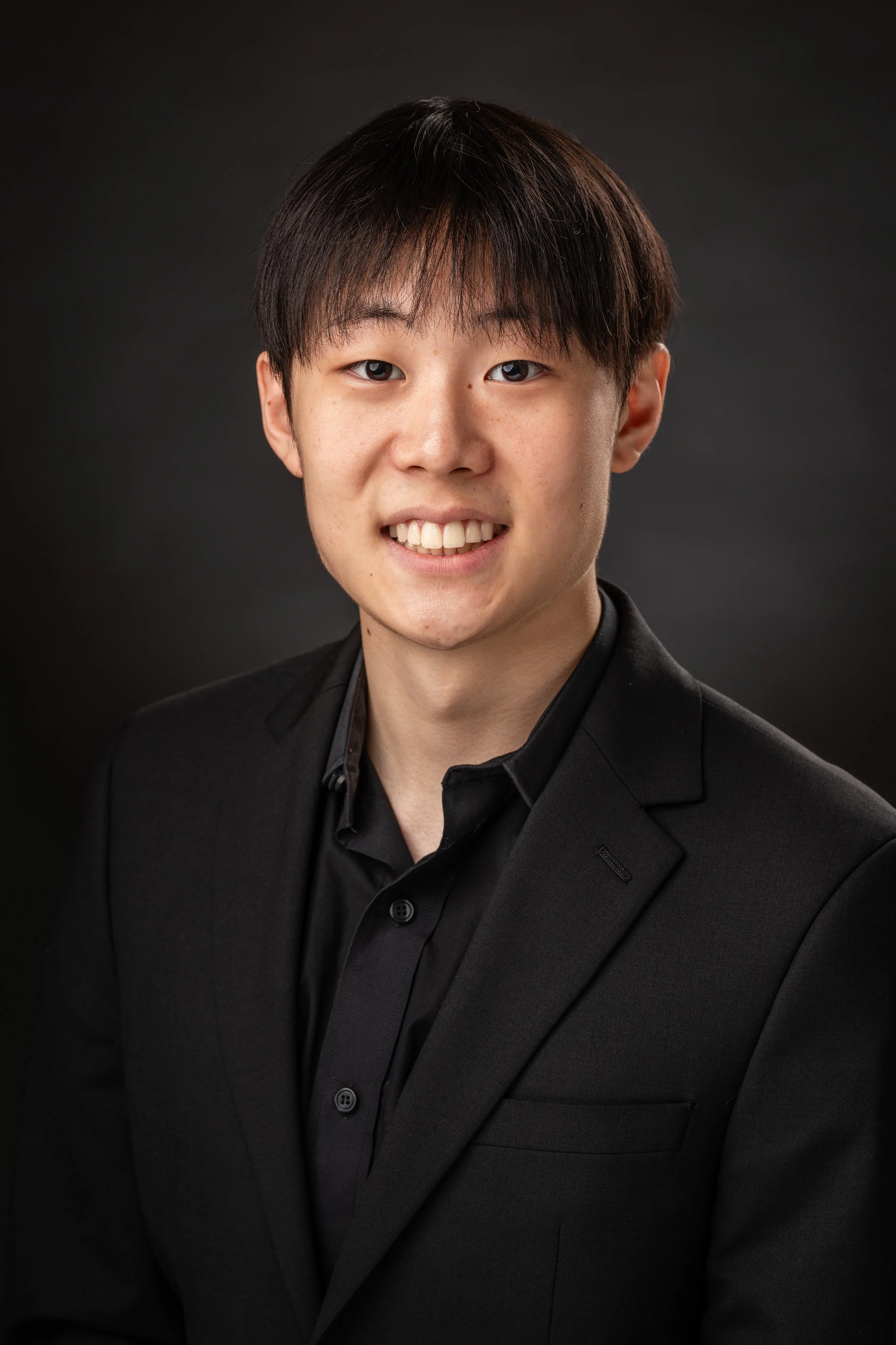 Young man in black suit and shirt smiling against black background.