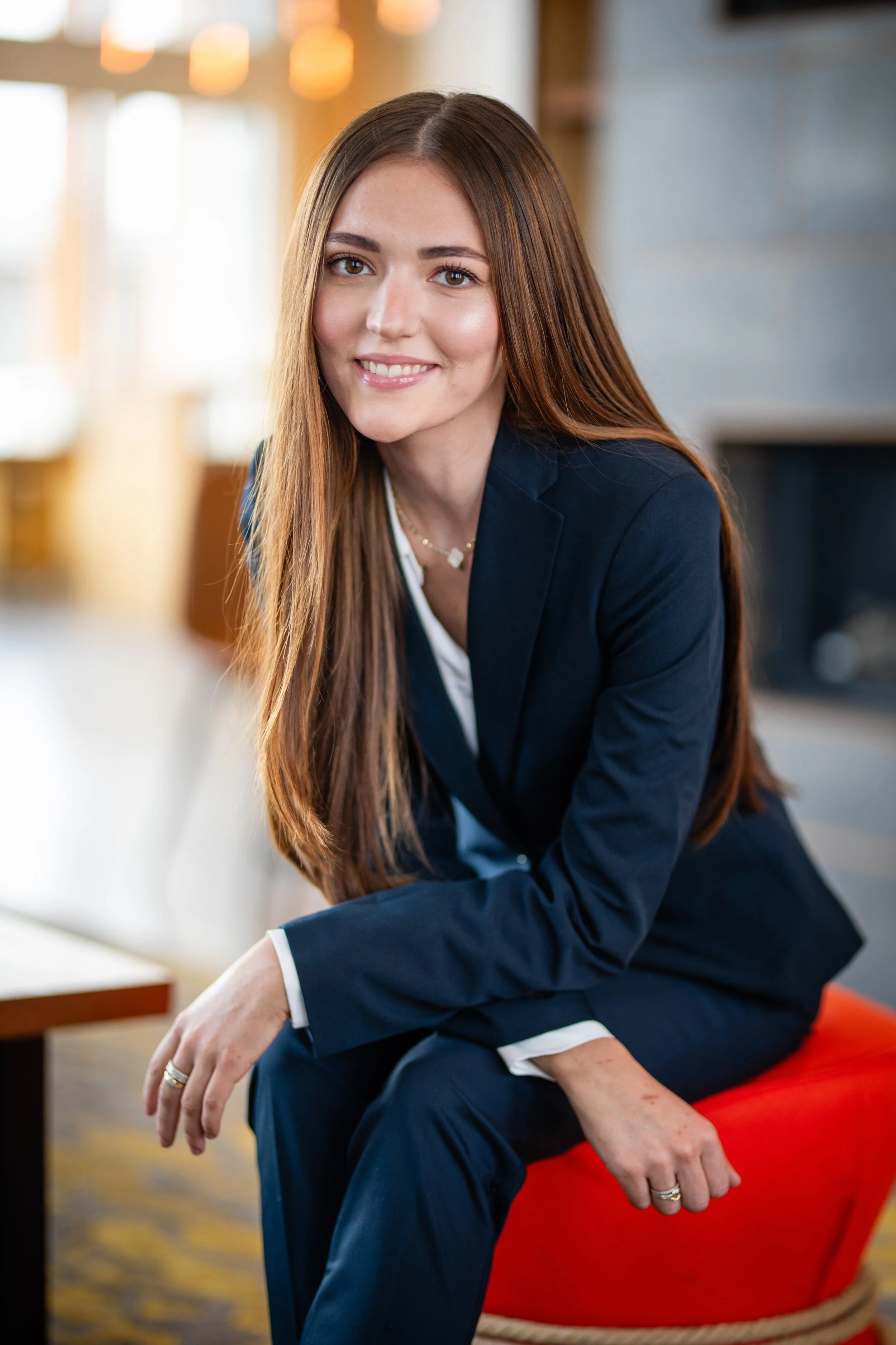 A woman with long brown hair wearing a navy blue suit, smiling while sitting on a red ottoman in a well-lit indoor setting.