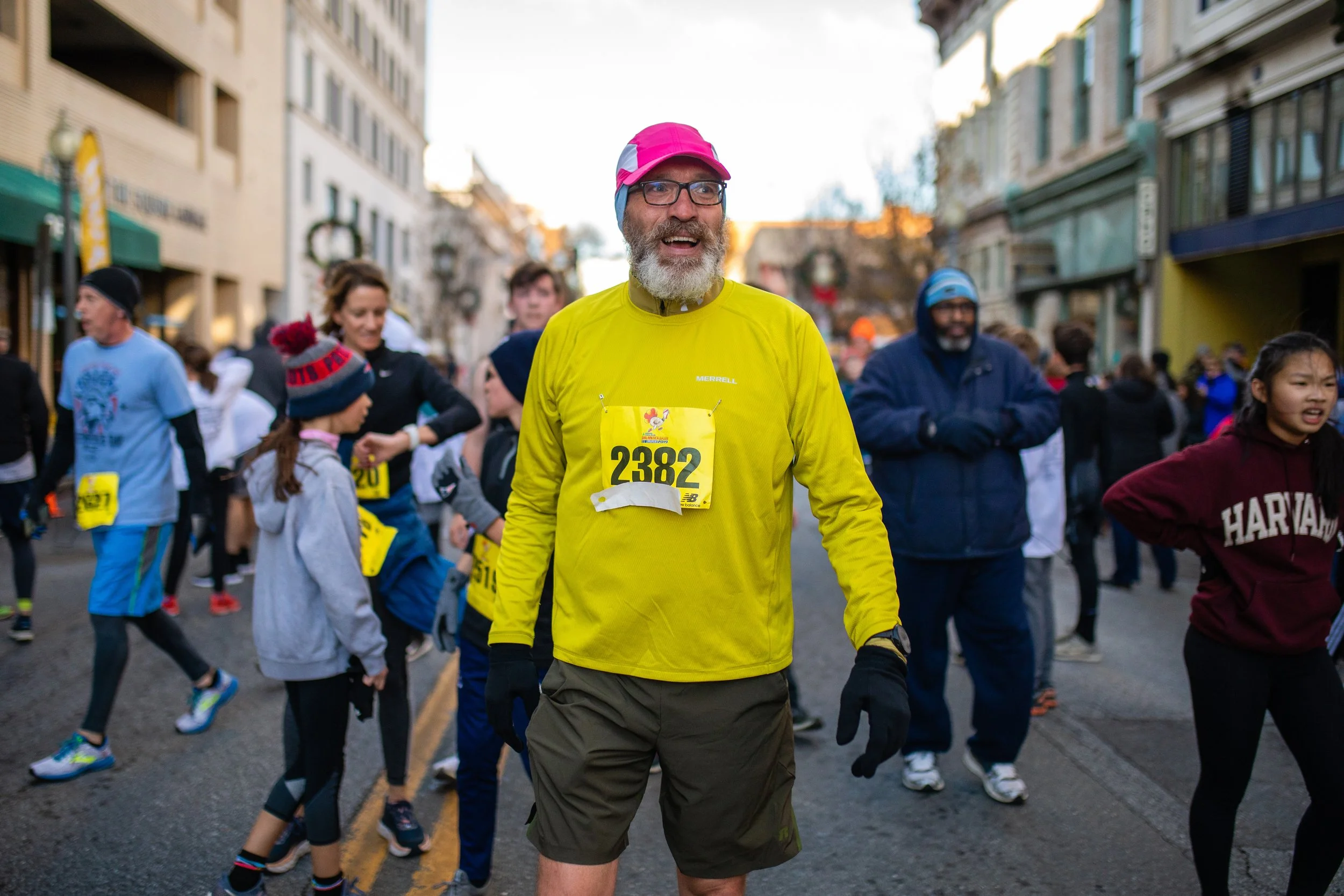 A man with glasses and a beard wearing a bright yellow shirt and a pink cap standing among runners at a street race. Many runners are dressed in athletic clothes, and buildings line the street.