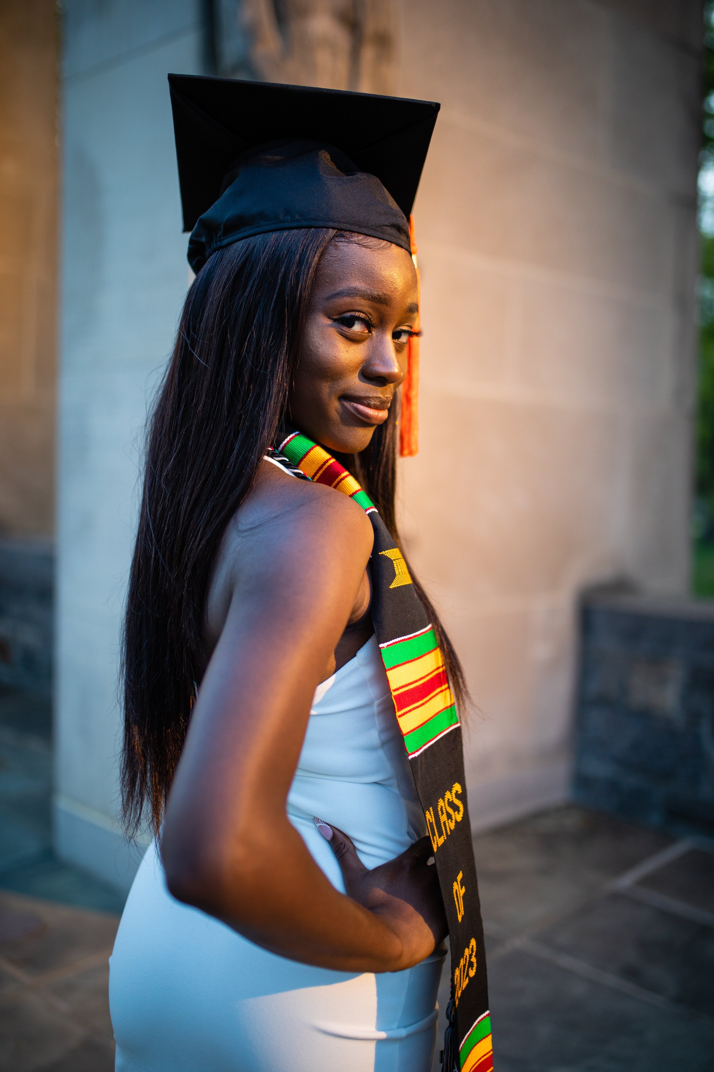 A young woman in a white dress wearing a graduation cap and a colorful kente cloth stole, smiling and posing outdoors.
