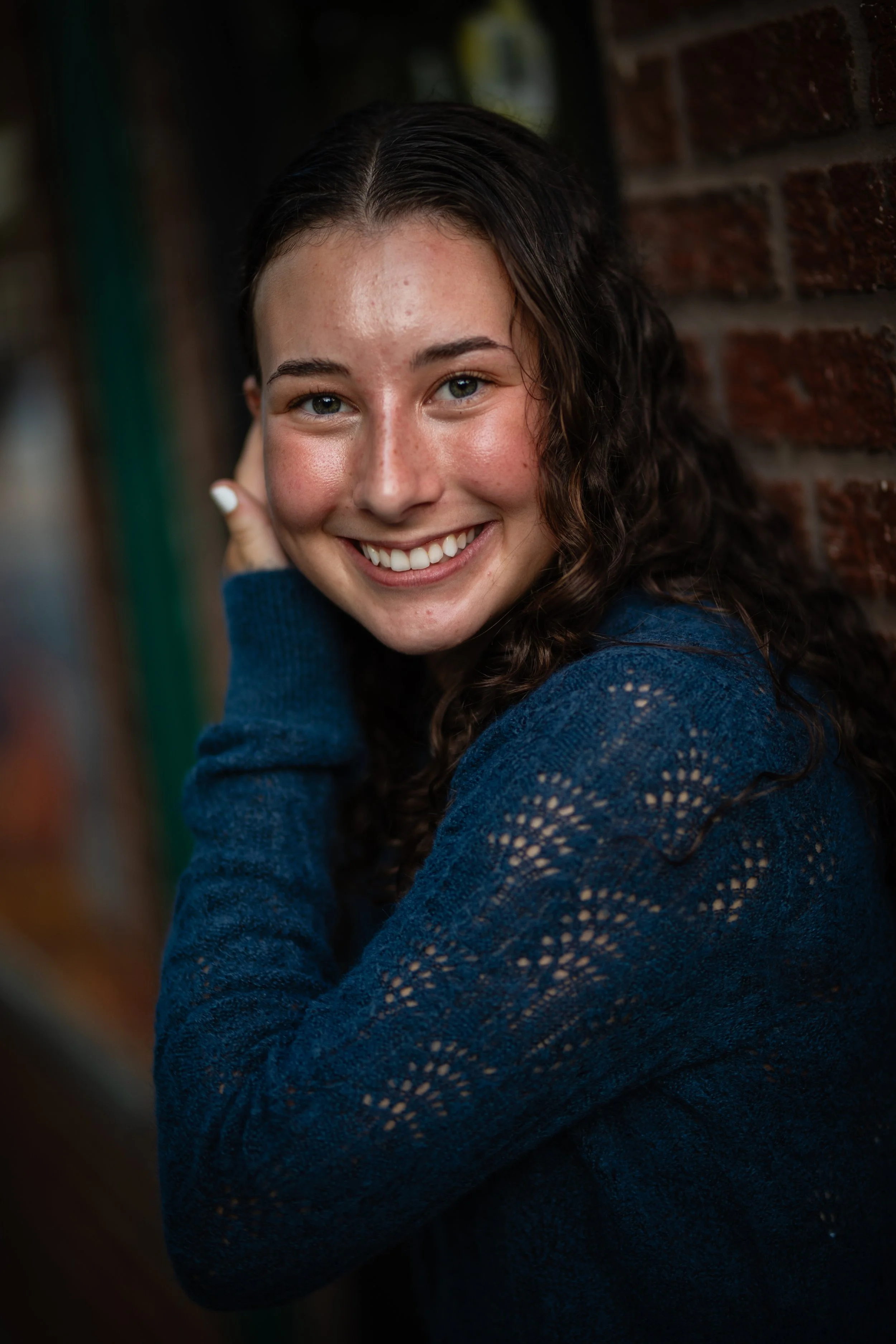 A young woman with curly brown hair, smiling, wearing a dark blue sweater, and standing against a brick wall.