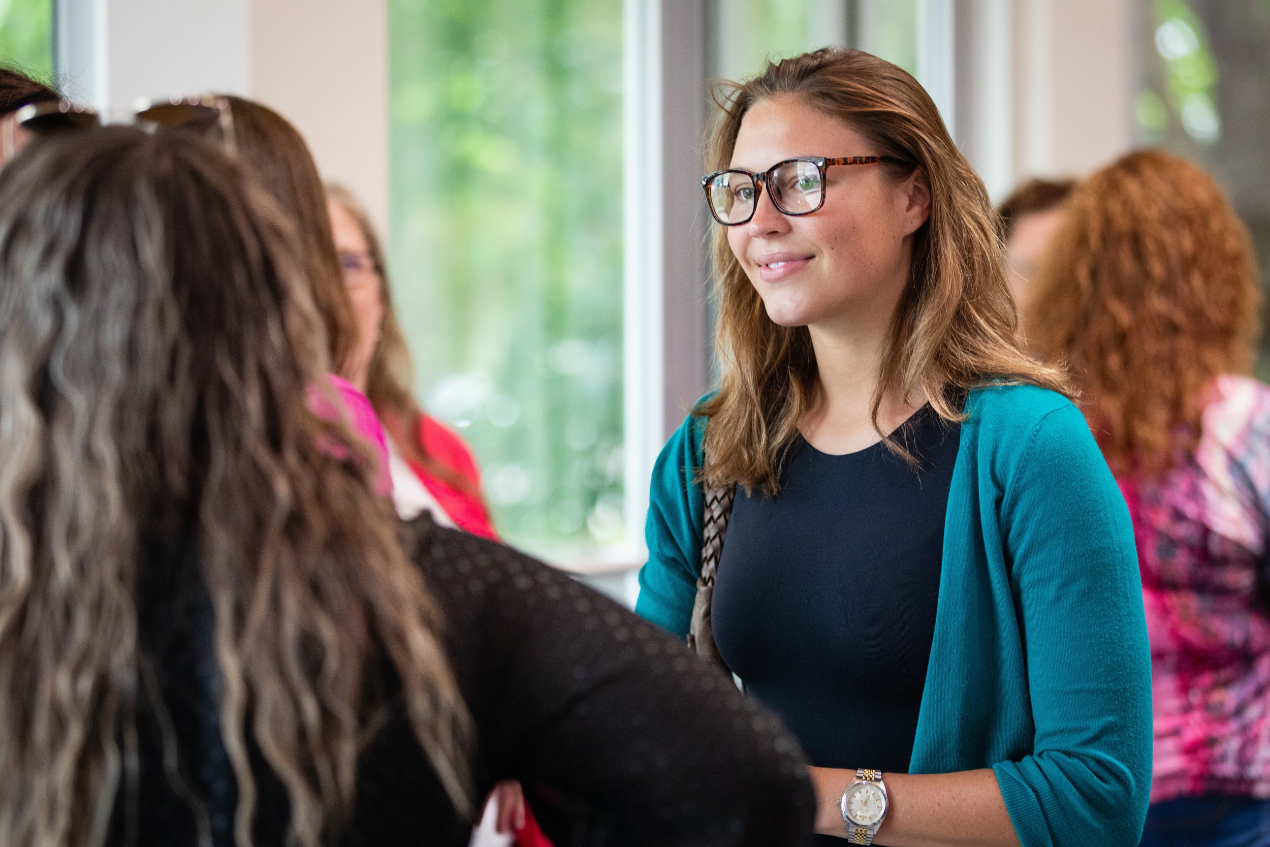 A woman with glasses and a blue cardigan smiling and talking to another woman with long curly brown hair at a social gathering indoors near a window.